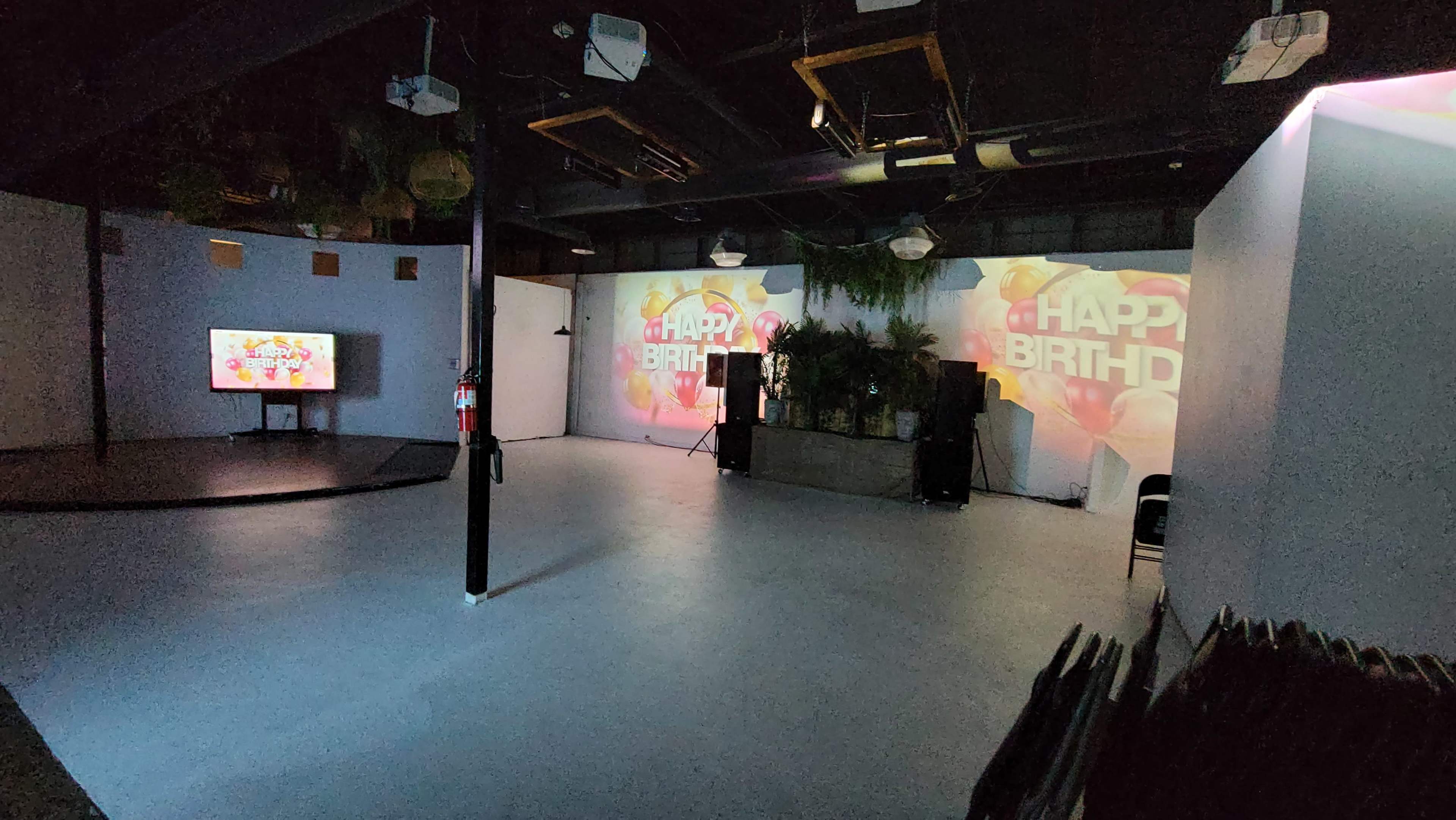 A spacious event room with a large "Happy Birthday" projection on the wall, a television, and sound equipment arranged near a planter.