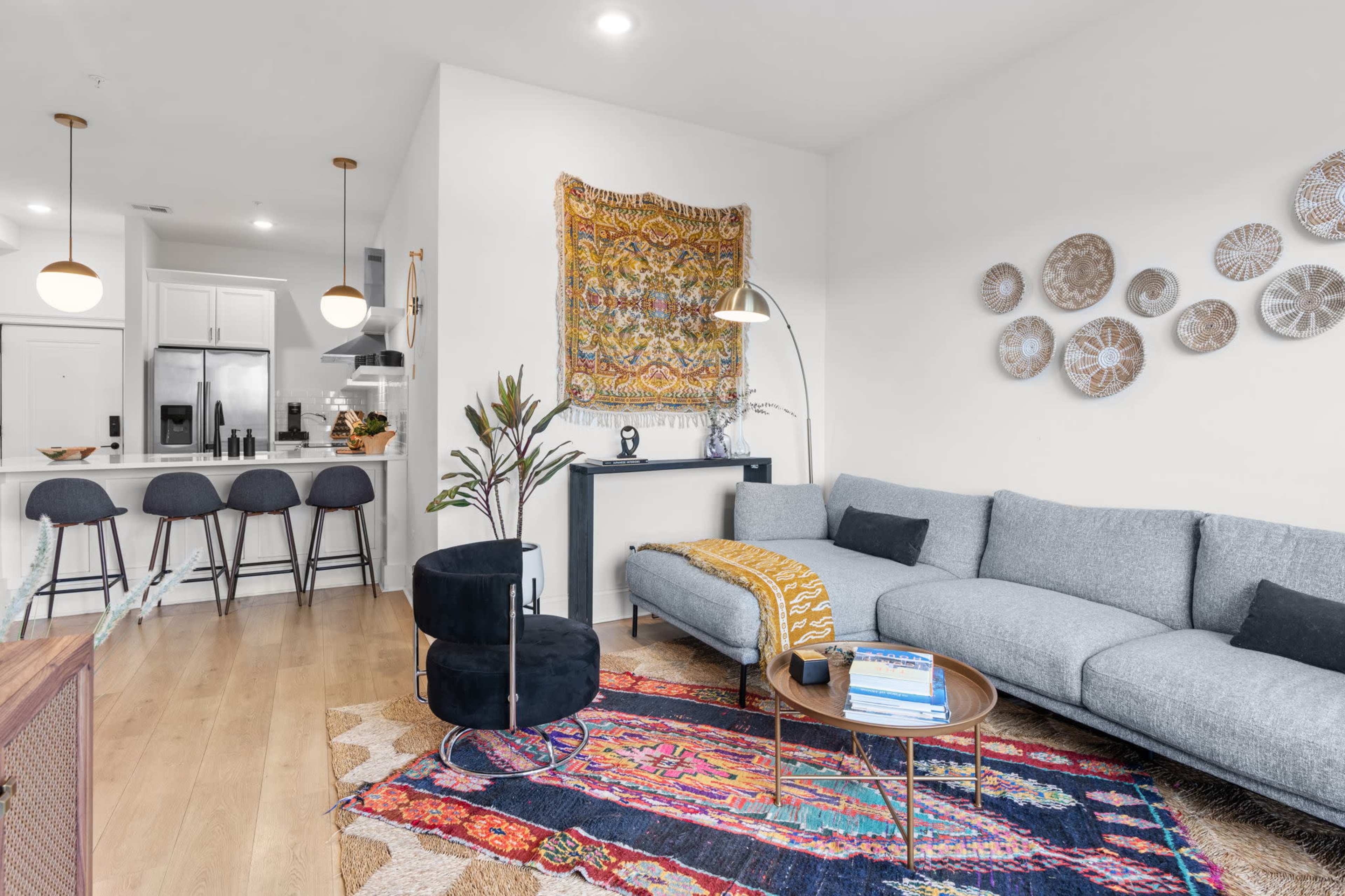 A modern living room with a gray sectional sofa, a round coffee table, decorative wall art, and a kitchen area in the background with bar stools.