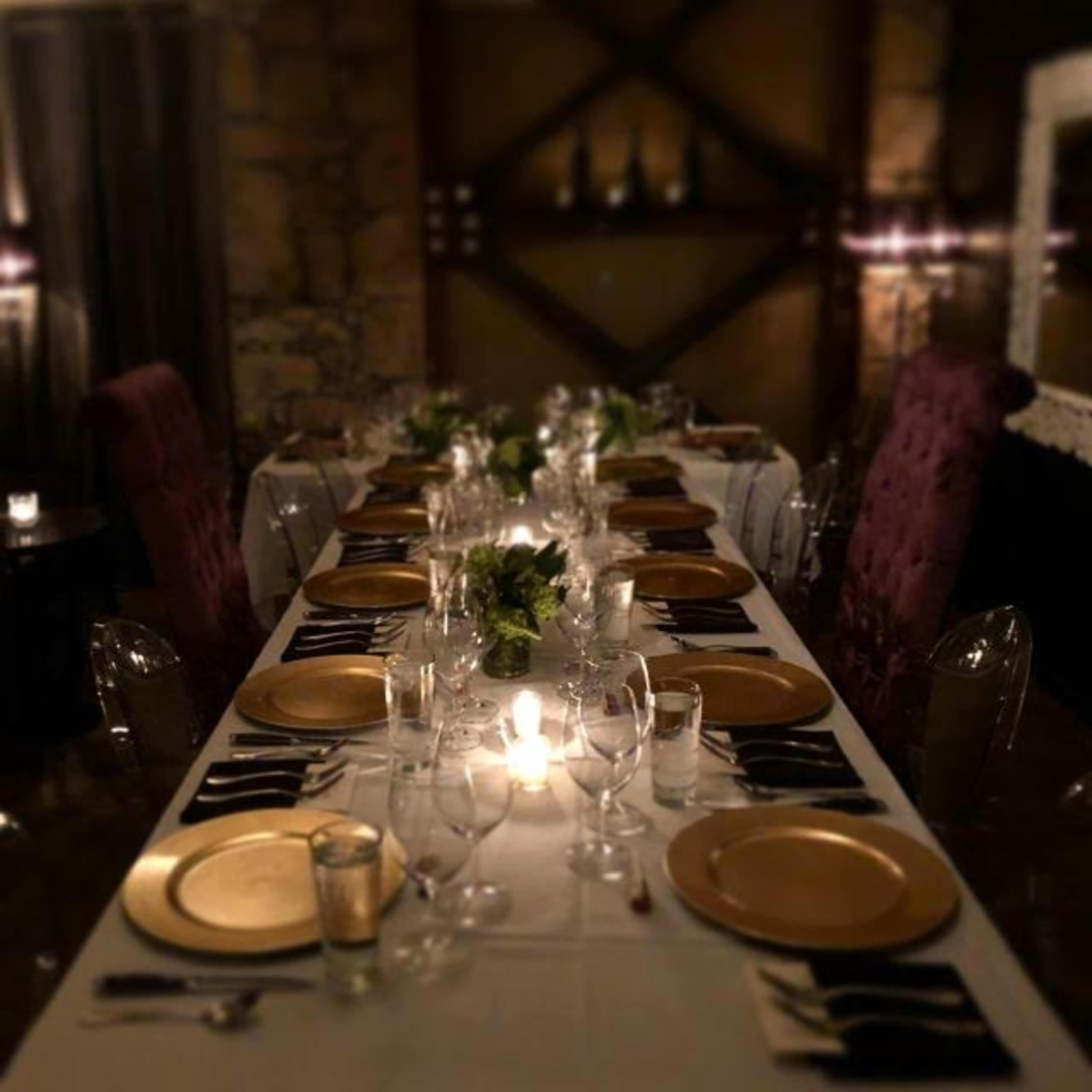 A long dining table is set with gold plates, glassware, and foliage in a dimly lit room.