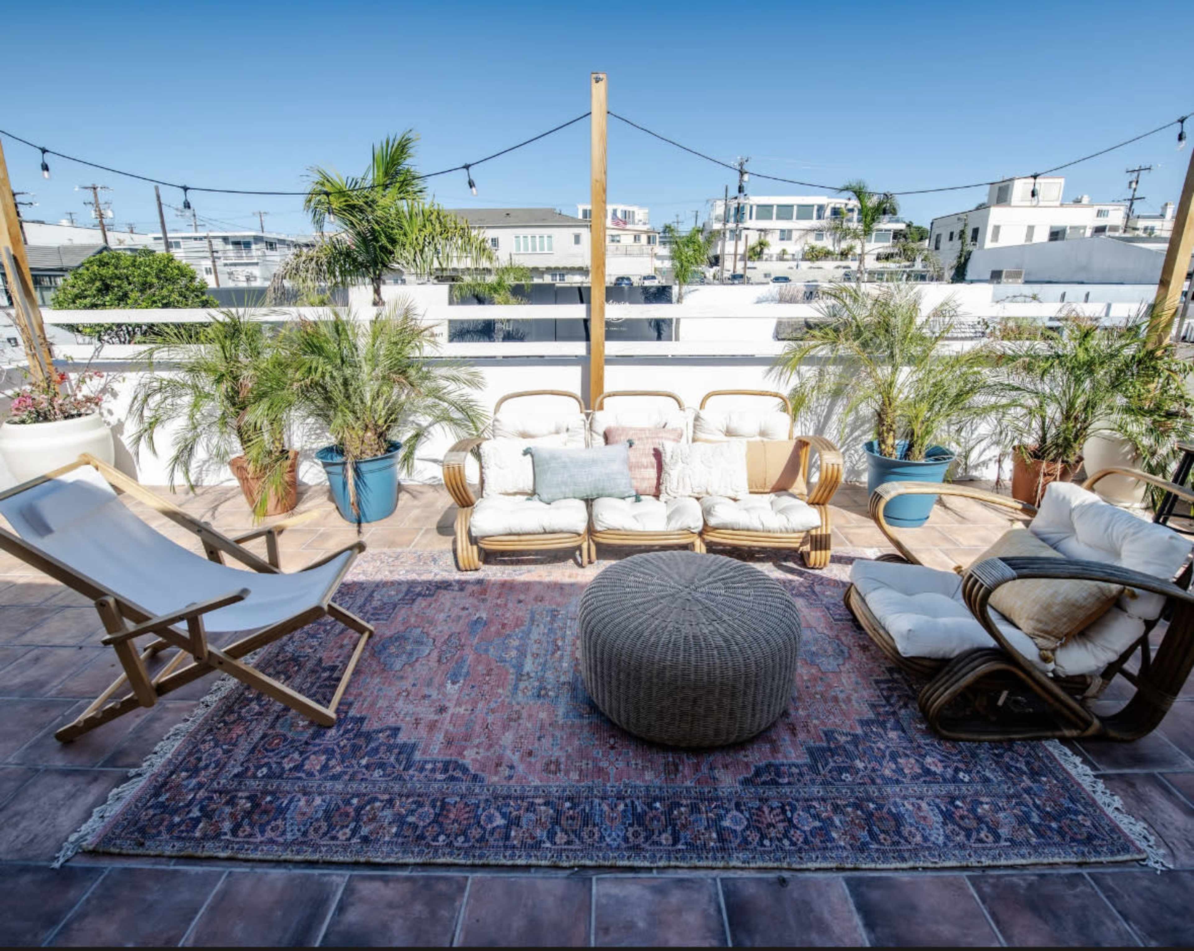 A rooftop patio features several seating arrangements, including a sofa and reclining chair, surrounded by potted plants and a large area rug.