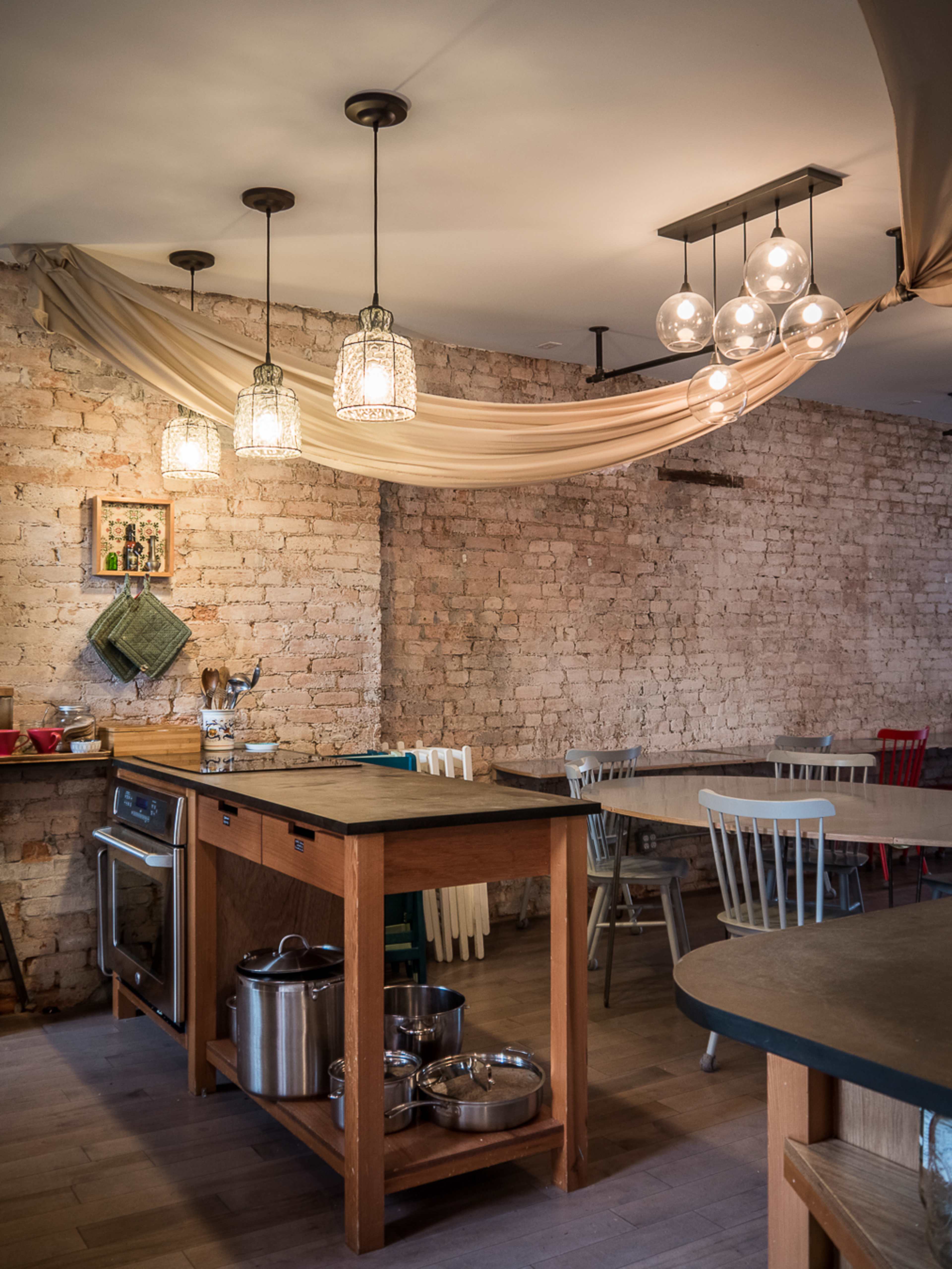 A kitchen area with wooden furniture, a cooking station, and several pendant lights hanging from the ceiling against a brick wall.