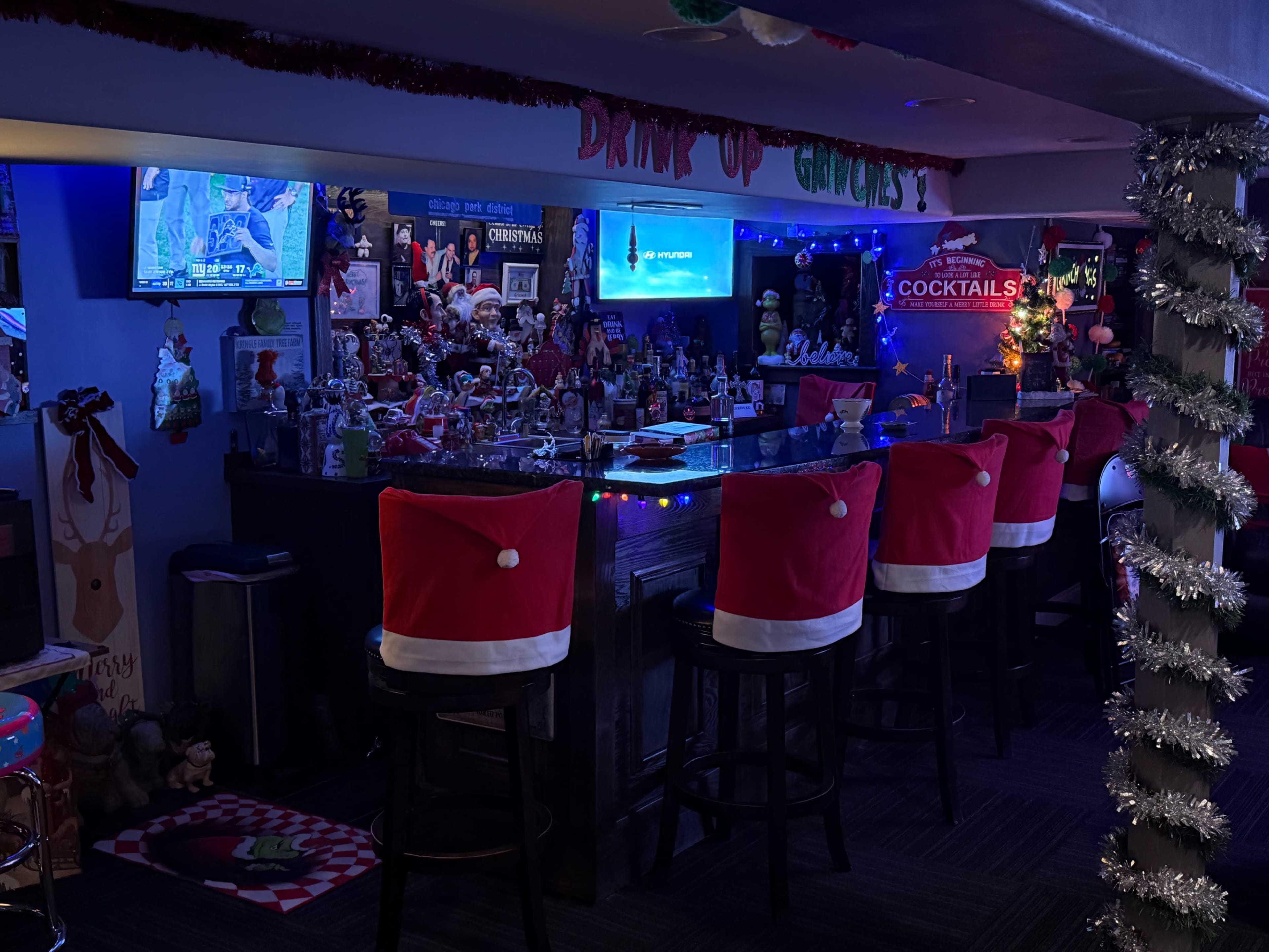 A festive bar decorated for Christmas features stools with red Santa hat covers and colorful lights illuminating various holiday decorations.