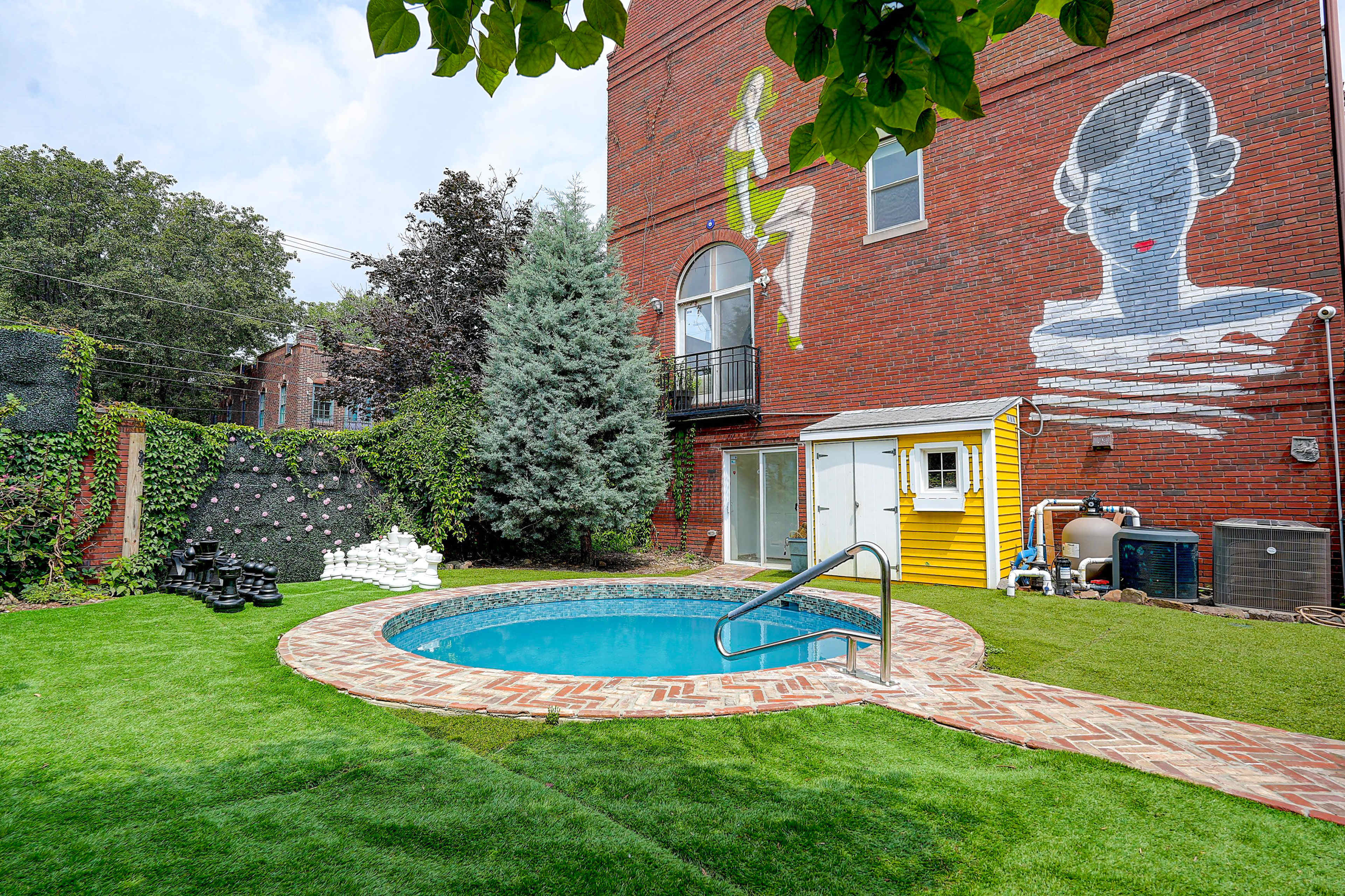 The scene features a small circular pool surrounded by a brick patio and a lush green lawn, with a mural of a woman on the brick wall of a building in the background.