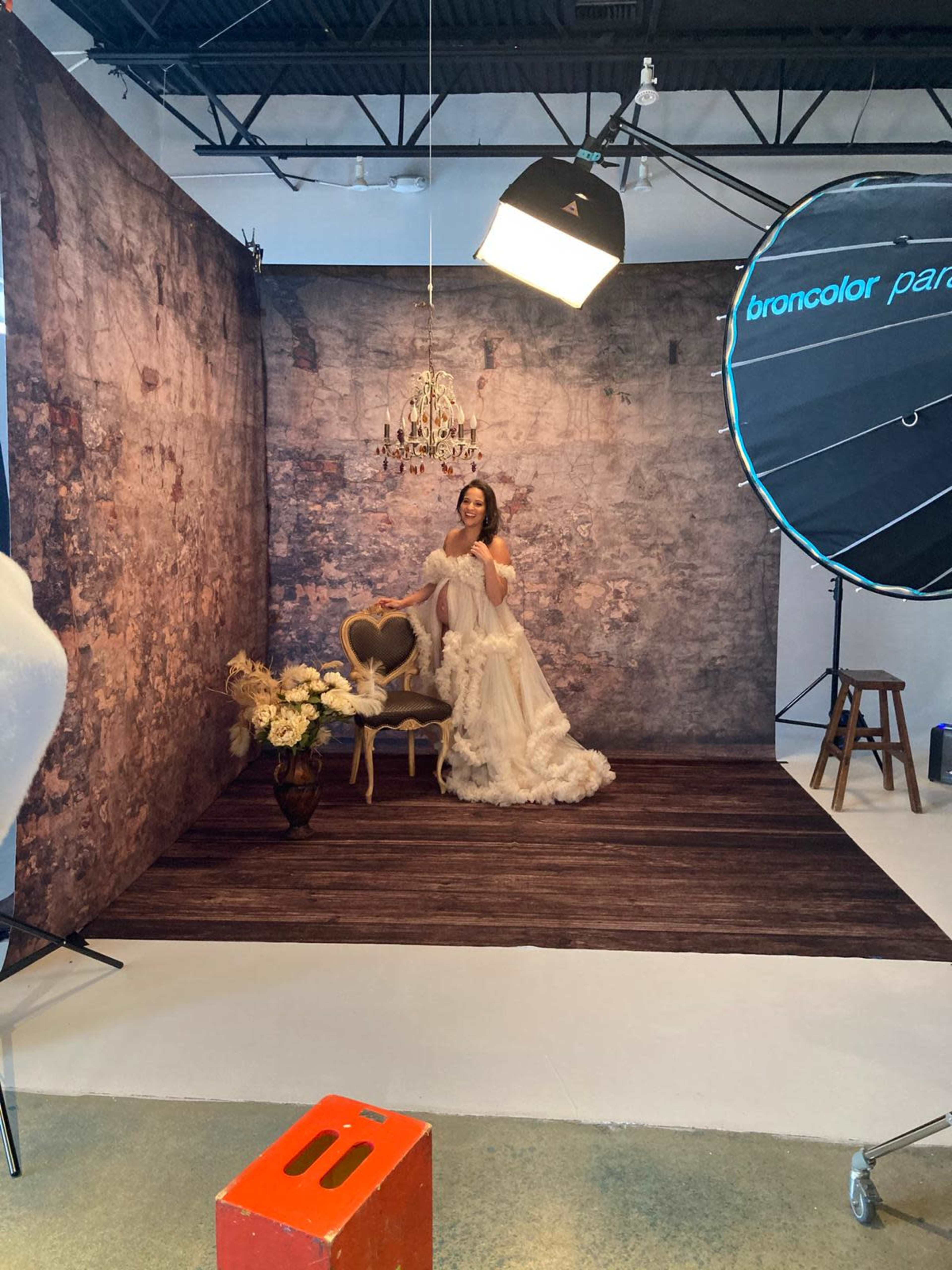 A woman in a white dress poses in a photography studio with a vintage-style chair and a chandelier in the background.