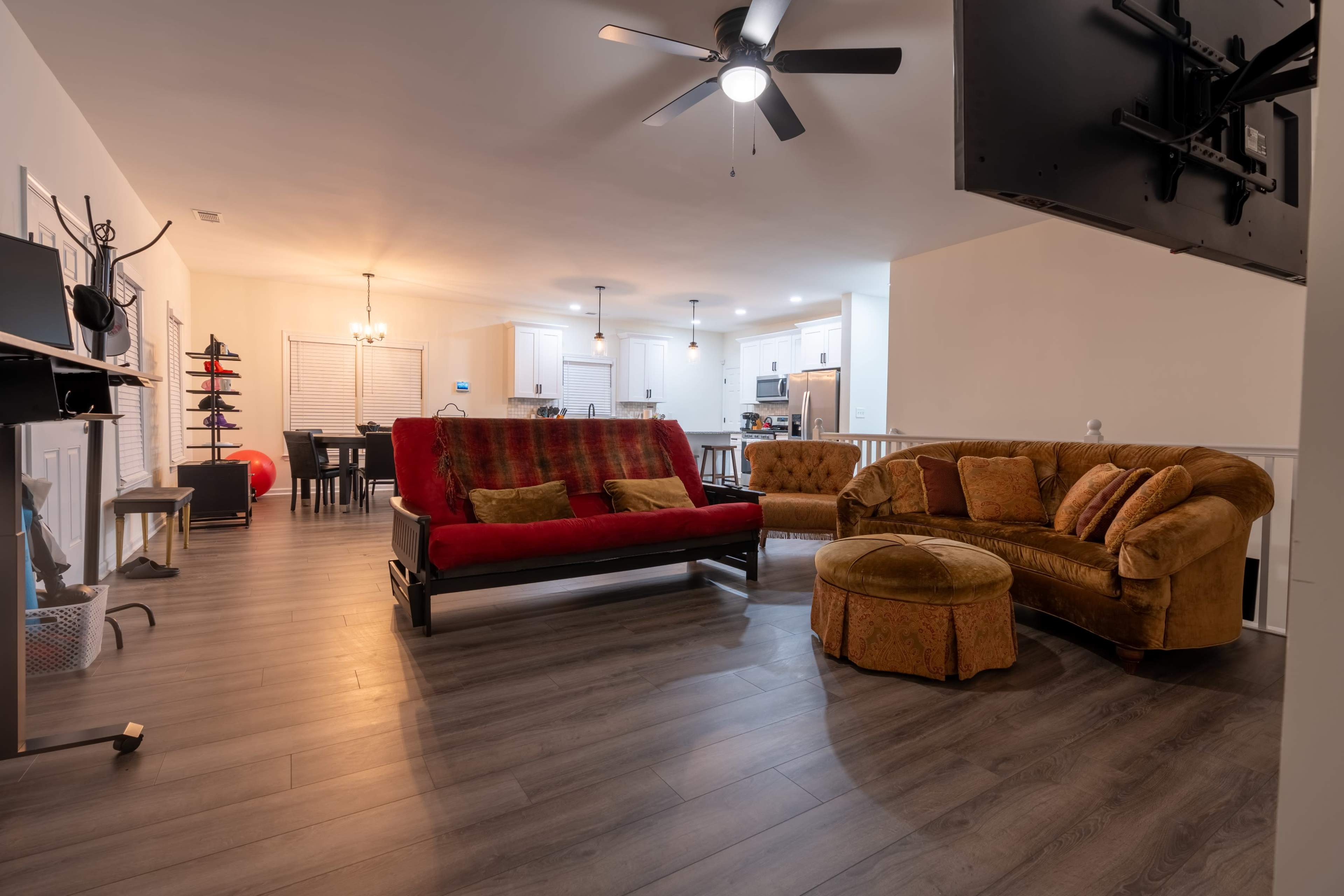 The image shows a spacious living area with a red sofa, a brown couch, and an ottoman, leading to a well-lit kitchen in the background.
