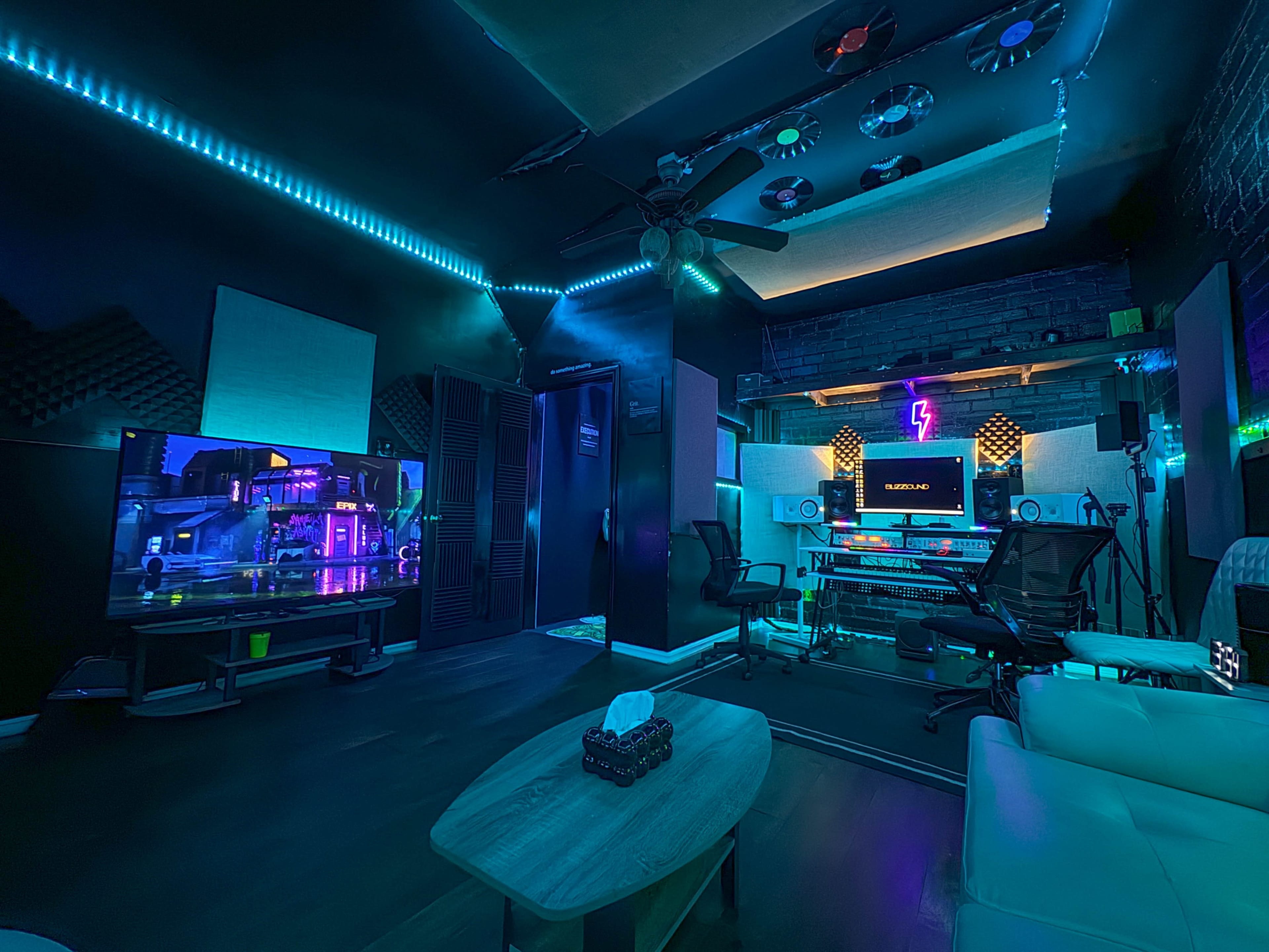 A modern music studio with neon lighting, a large monitor, and audio equipment arranged around a central coffee table and seating area.