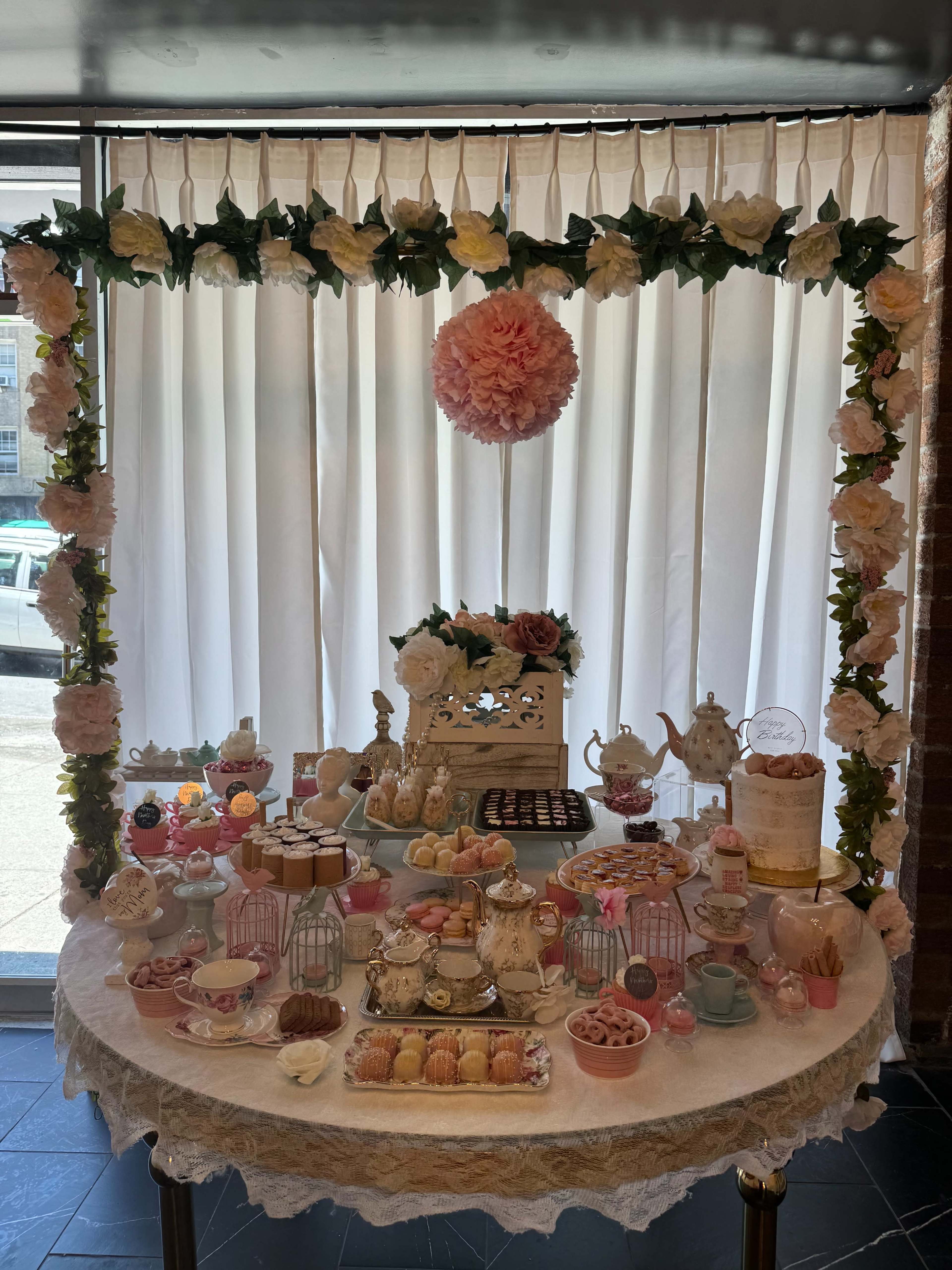 A table is adorned with an array of delicate pastries and a decorative tea set, set against a backdrop of draped white curtains and floral accents.