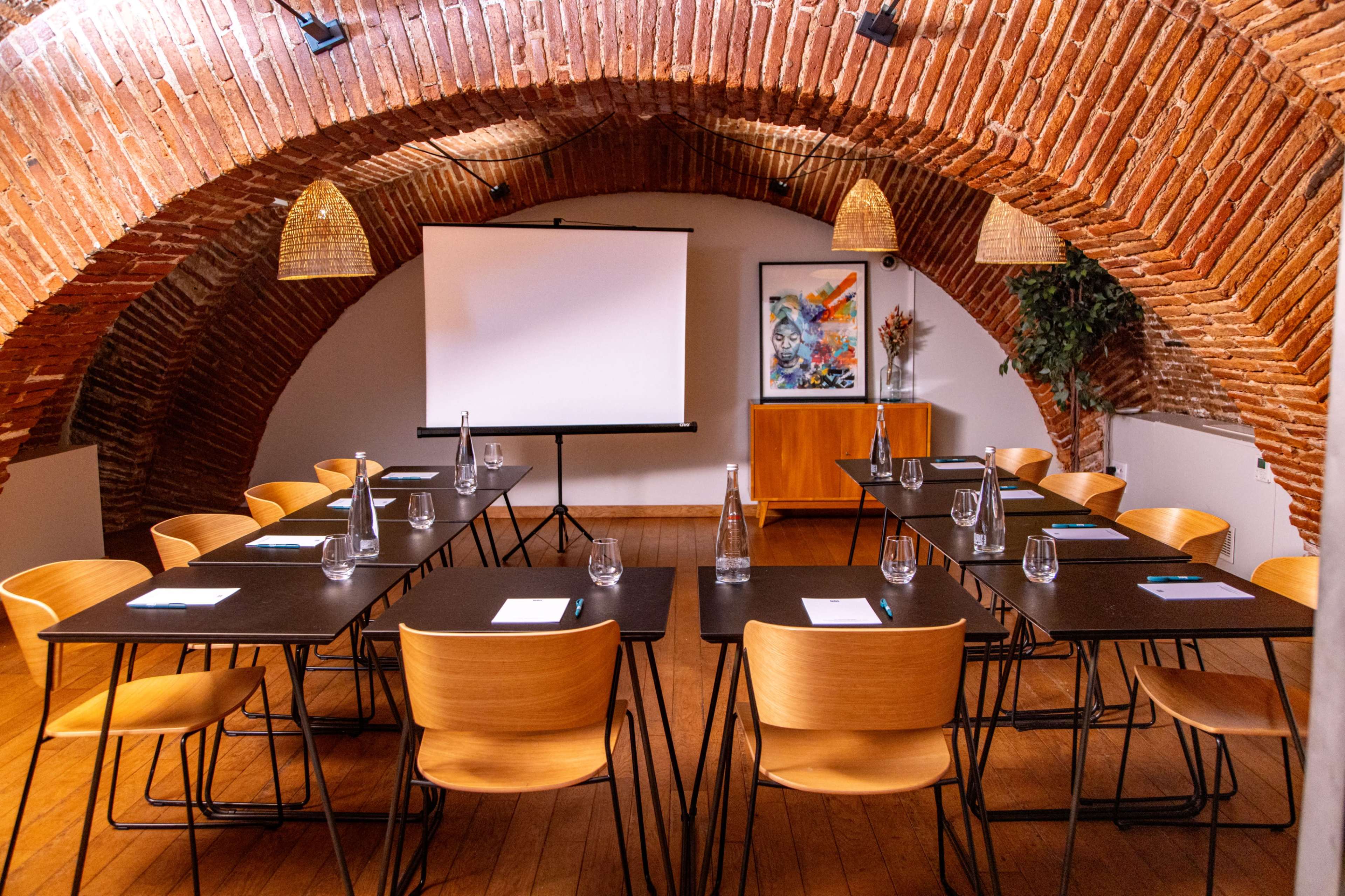 The image shows a conference room with a brick arch ceiling, featuring a central projection screen, wooden tables arranged in a U-shape, and chairs positioned around the tables.