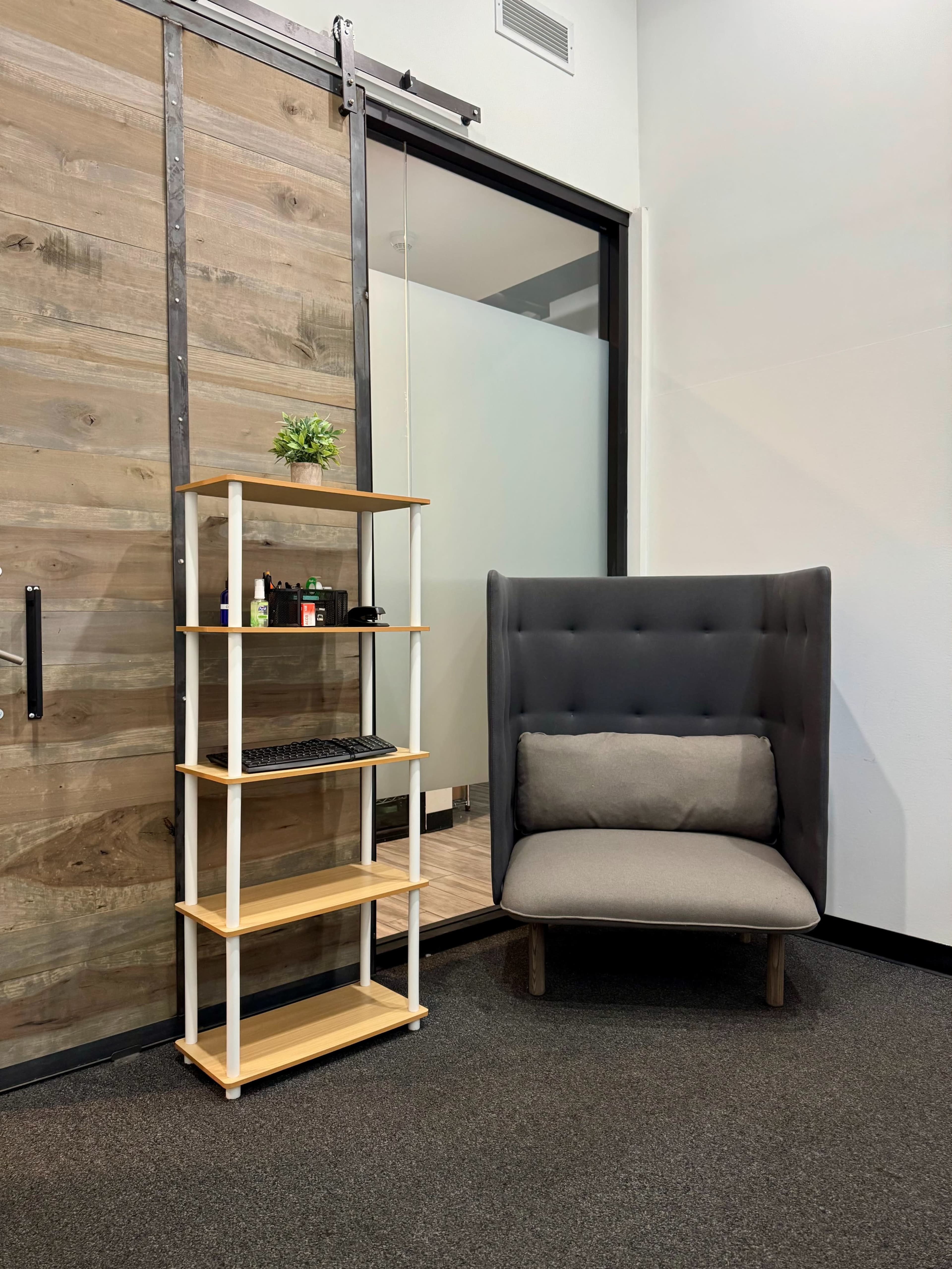 The image features a corner of a modern office space with a grey upholstered chair, a wooden shelving unit, and a sliding glass door.
