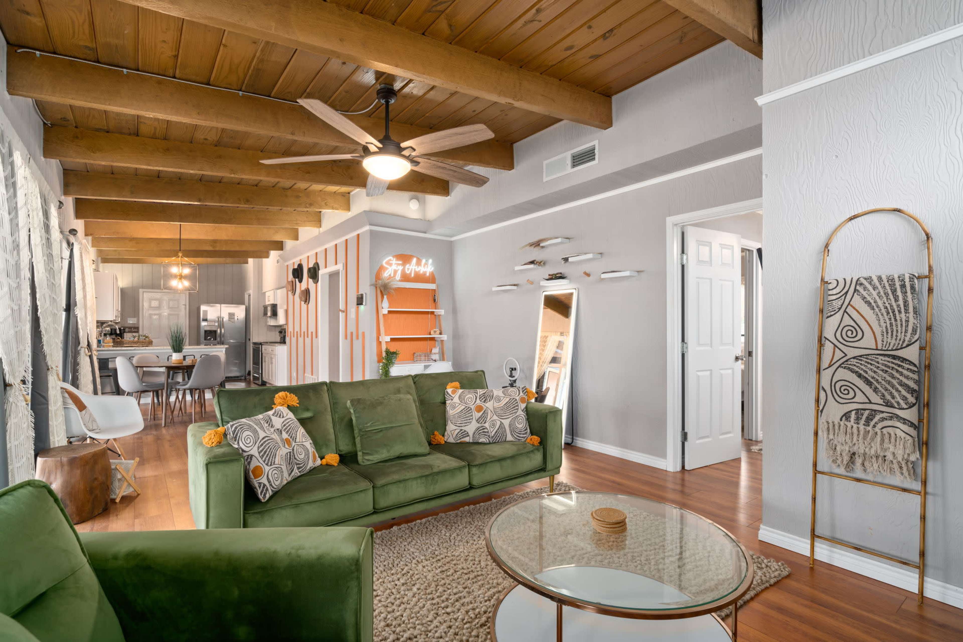 A living room with wooden beams on the ceiling, a green sofa adorned with decorative pillows, a glass coffee table, and an open layout leading to a kitchen.
