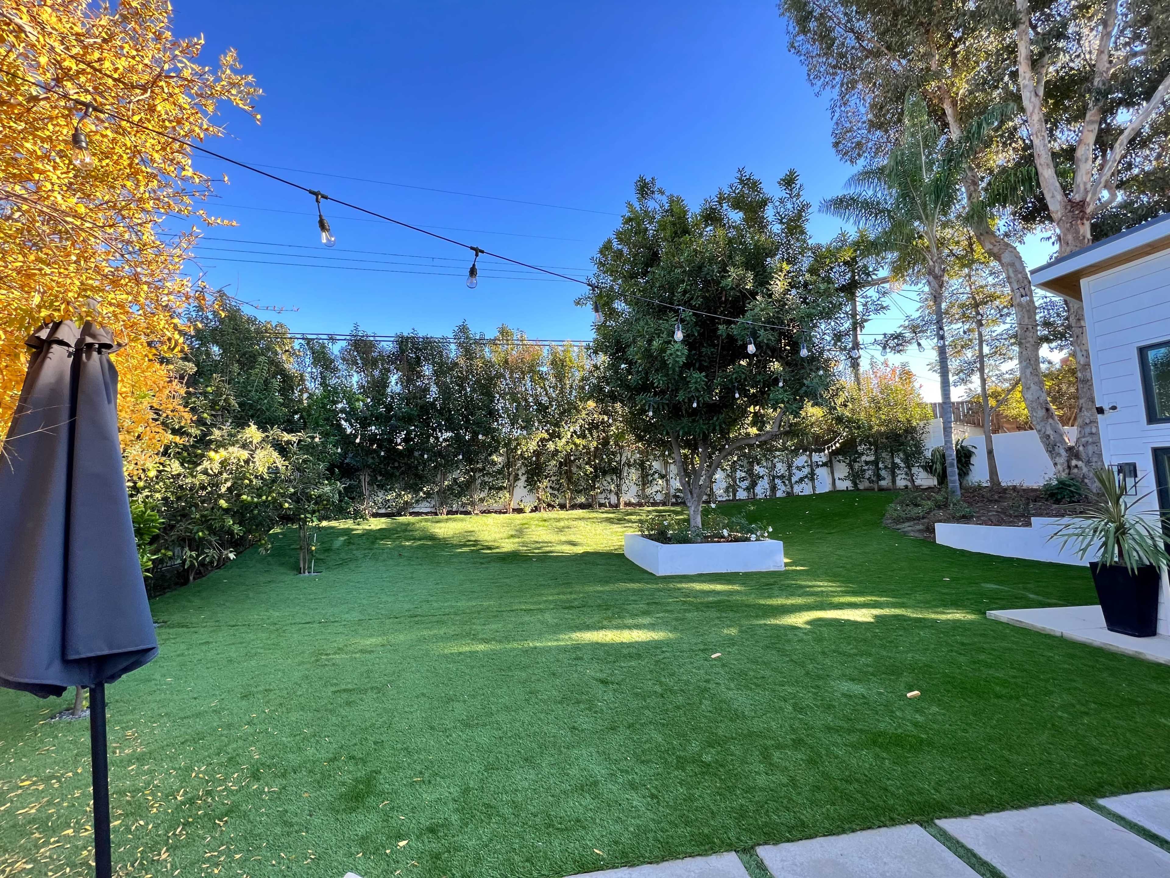 A well-maintained backyard with artificial turf, garden beds, and trees, illuminated by string lights overhead.