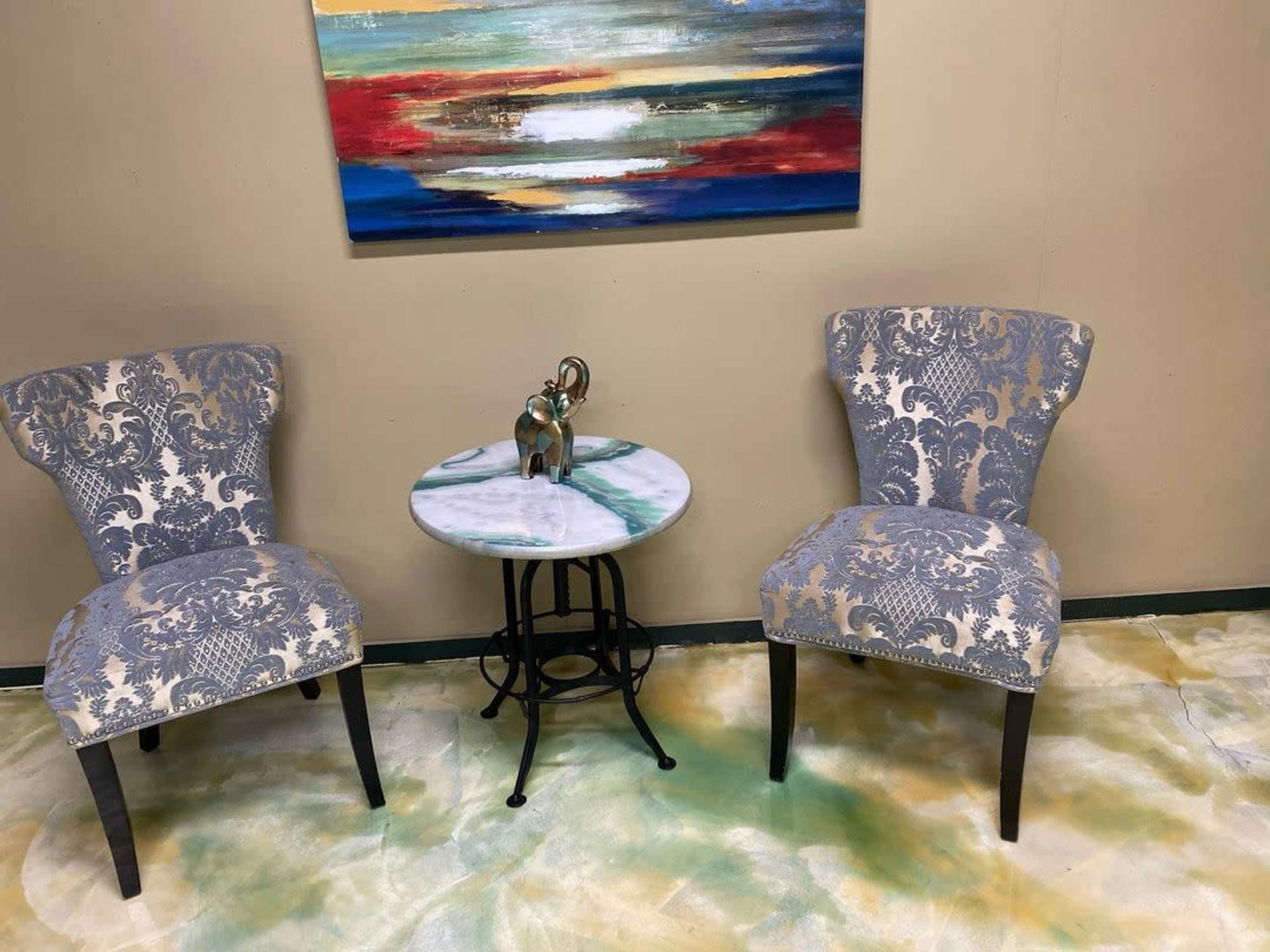 Two patterned chairs are positioned around a small round table with a green and white surface, set against a backdrop of a colorful abstract painting.