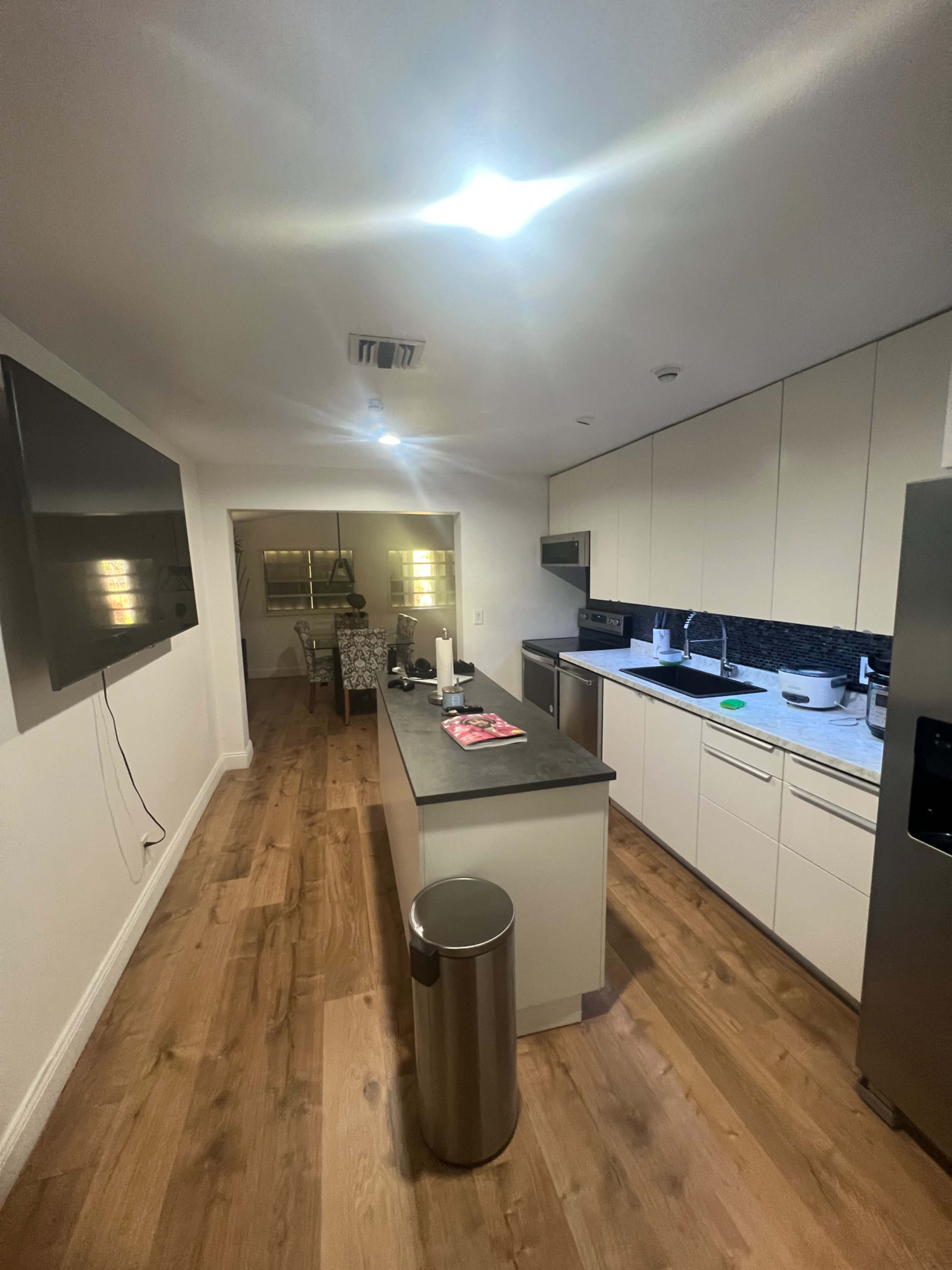 A modern kitchen with a central island, equipped with appliances, a television mounted on the wall, and wooden flooring.