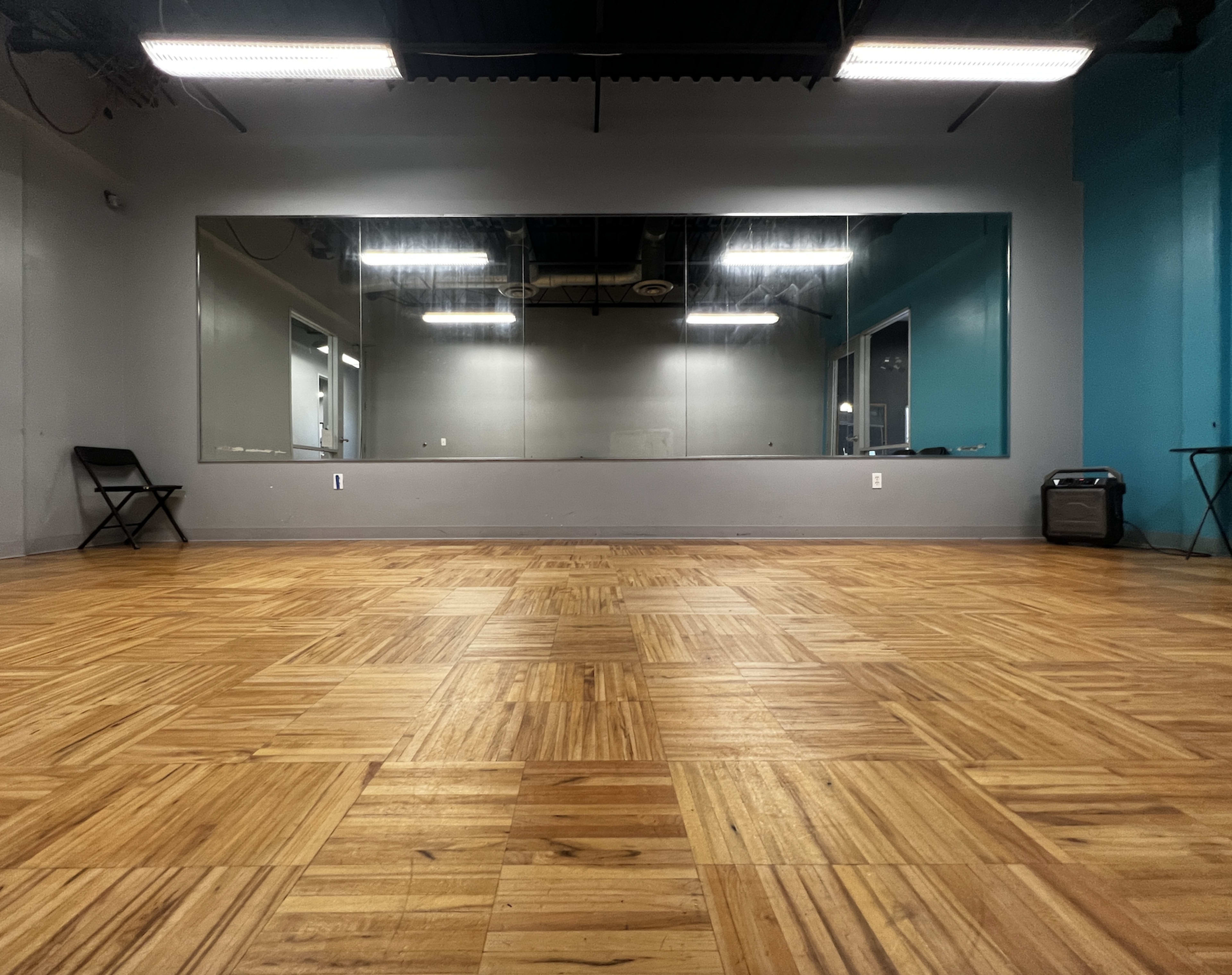 The image shows a spacious room with polished wooden flooring, large mirrors on one wall, and a blue accent wall opposite the mirrors.