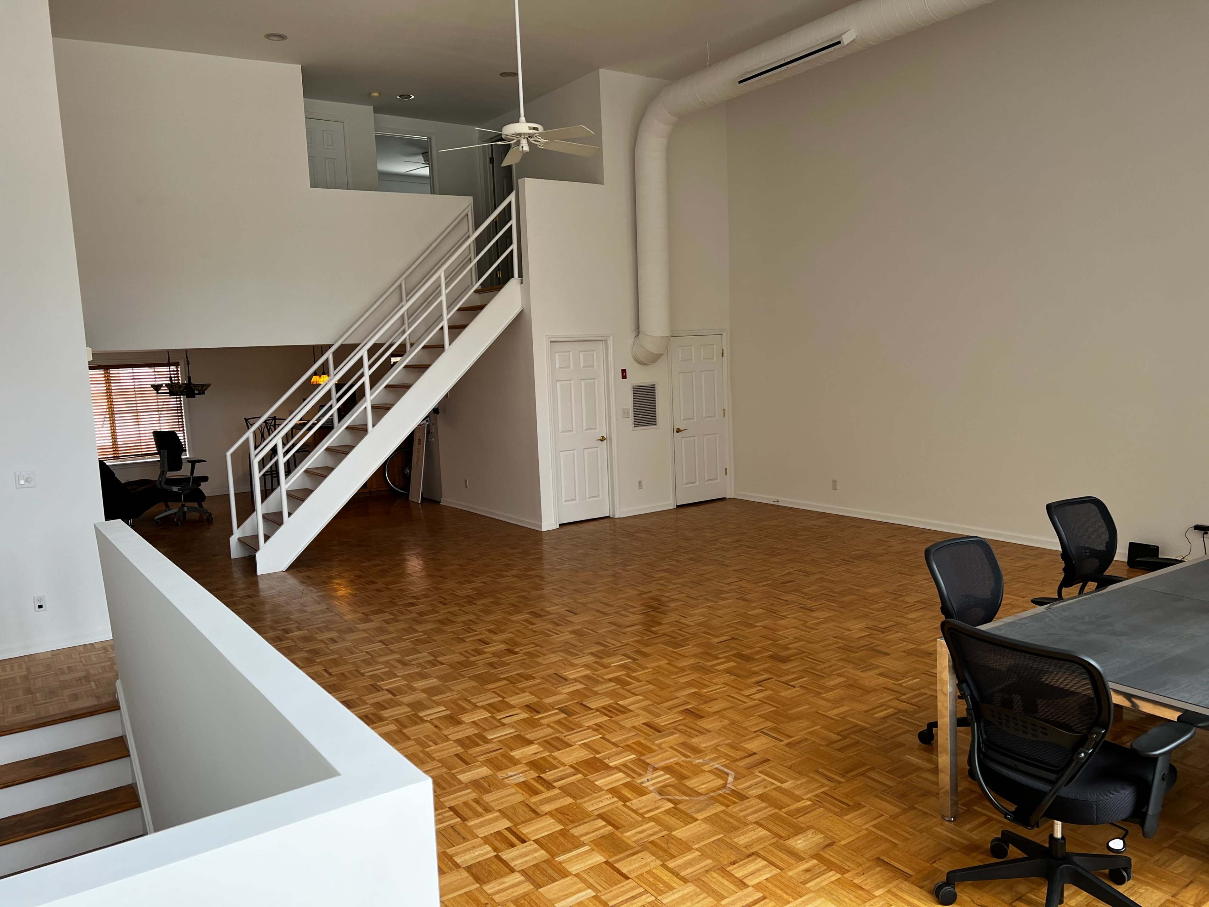 A spacious, open-plan room featuring wooden flooring, a staircase leading to a loft area, and a few office chairs arranged around a table.