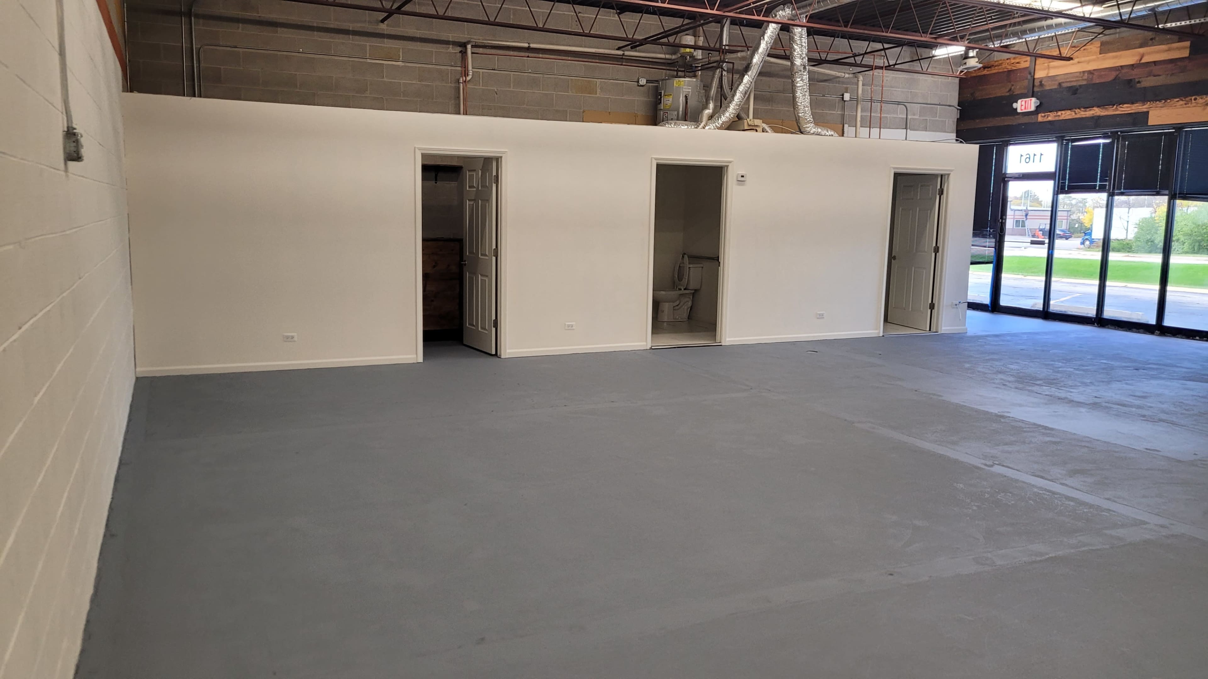 An empty commercial space with gray flooring and three doors leading to separate rooms along one wall, and large windows allowing natural light in.