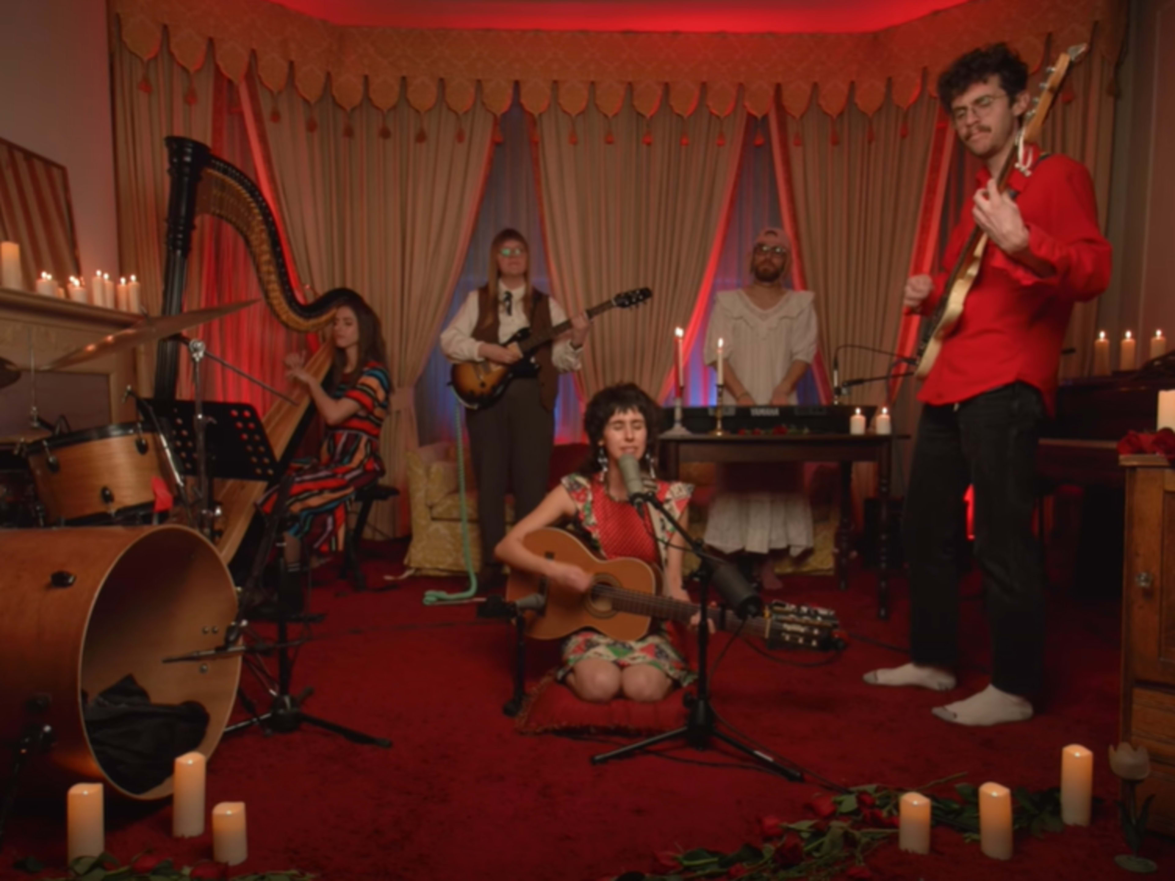 A band performs in a warmly lit room adorned with candles and red decor, featuring a harp, guitar, and drums.