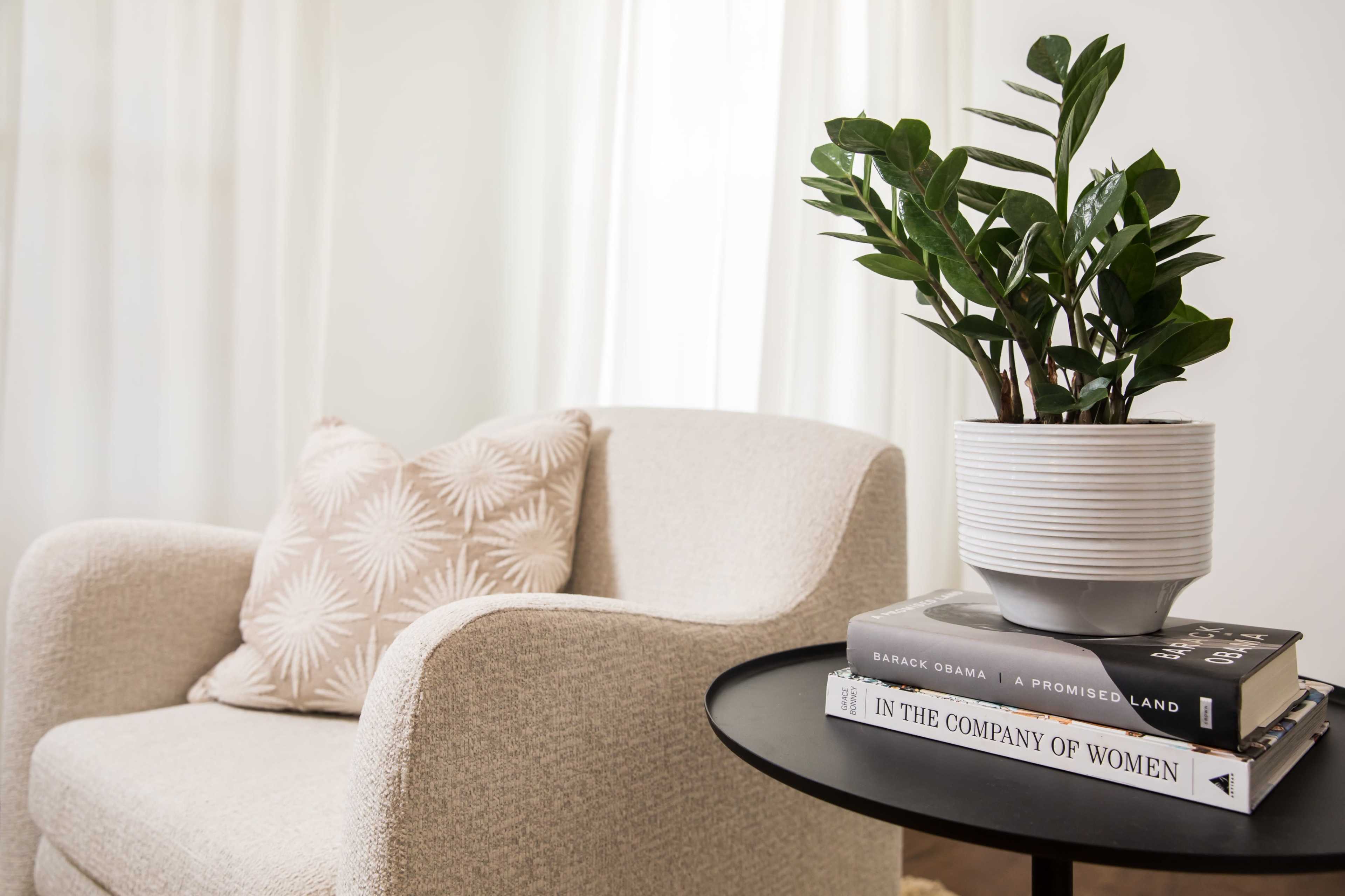 A light-colored chair with a patterned pillow sits beside a round table displaying a stack of books and a potted plant.