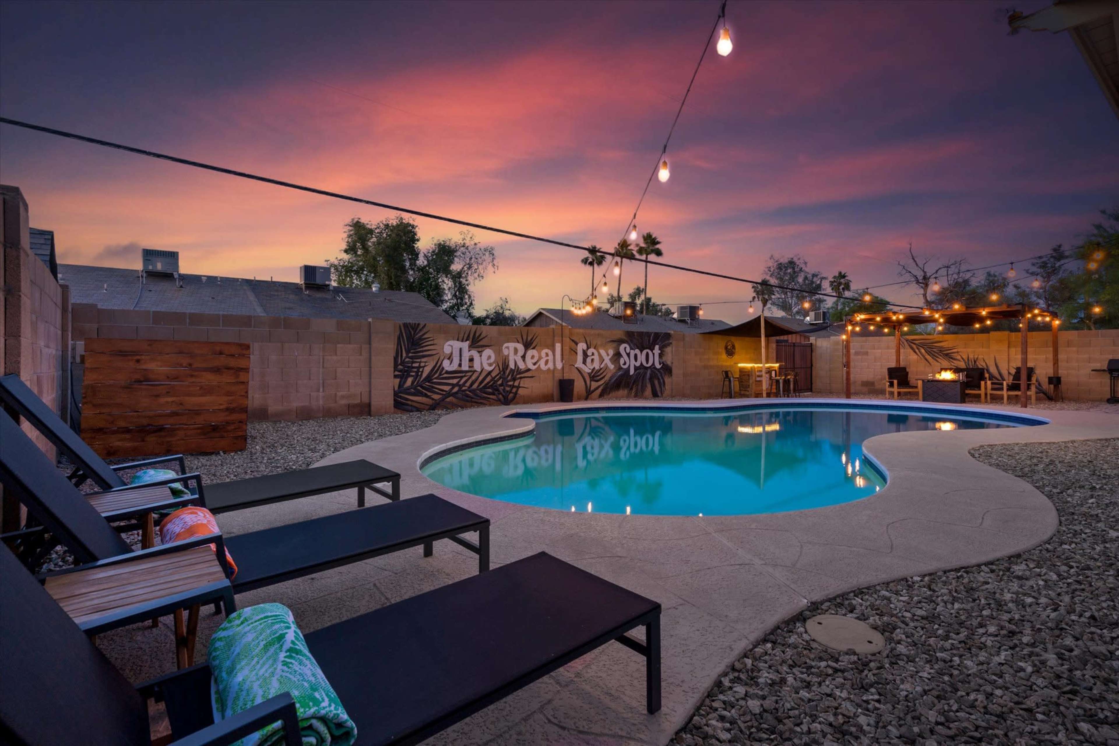 A swimming pool surrounded by lounge chairs and outdoor lighting, set against a colorful sunset sky.