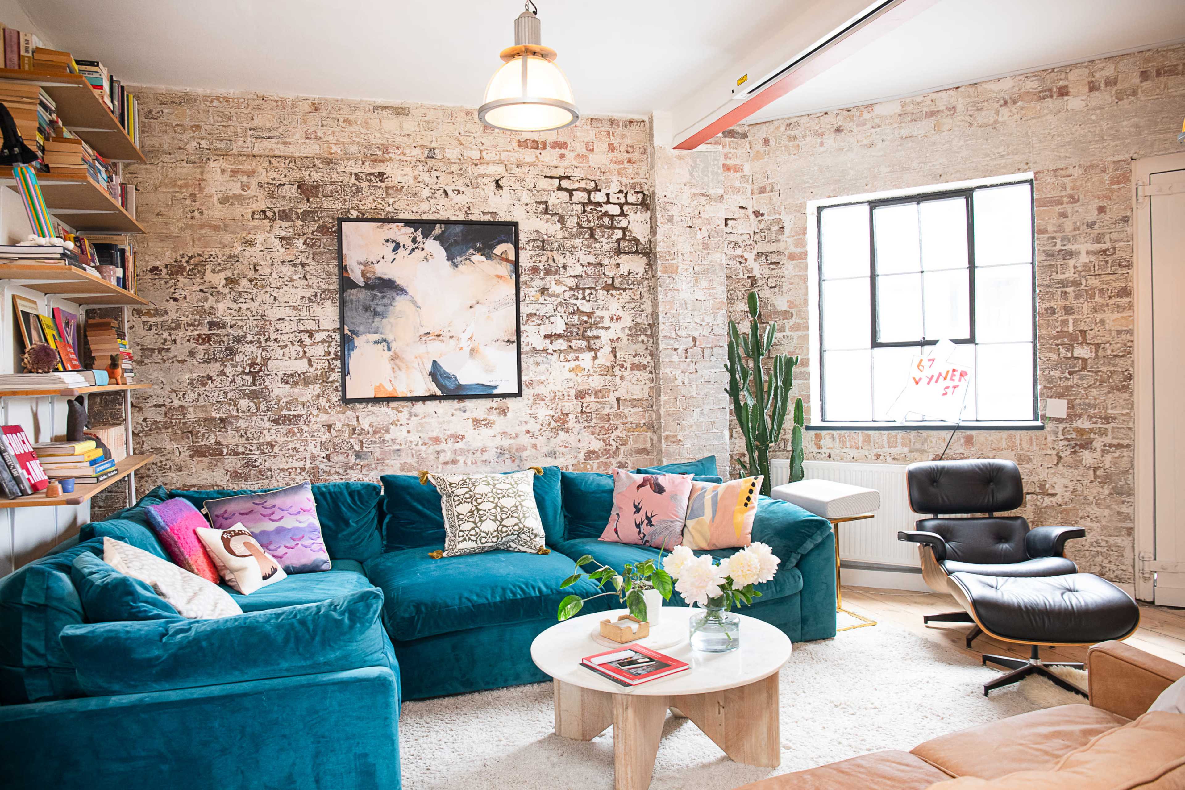 The image shows a cozy living room with exposed brick walls, a teal sofa, a round wooden coffee table, and a large window letting in natural light.