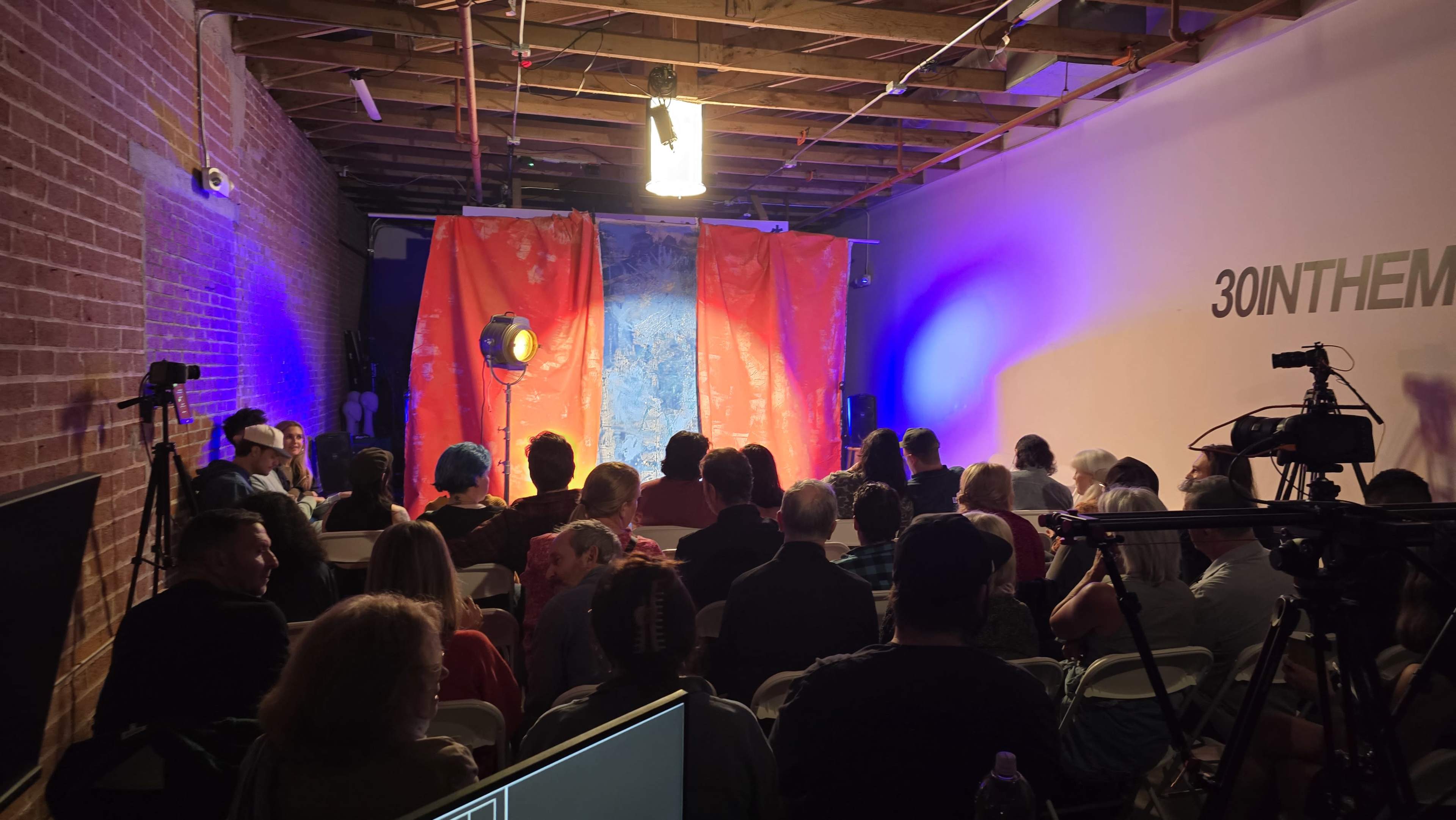 A crowd of people is seated in a room facing a brightly lit stage draped with colorful fabric.