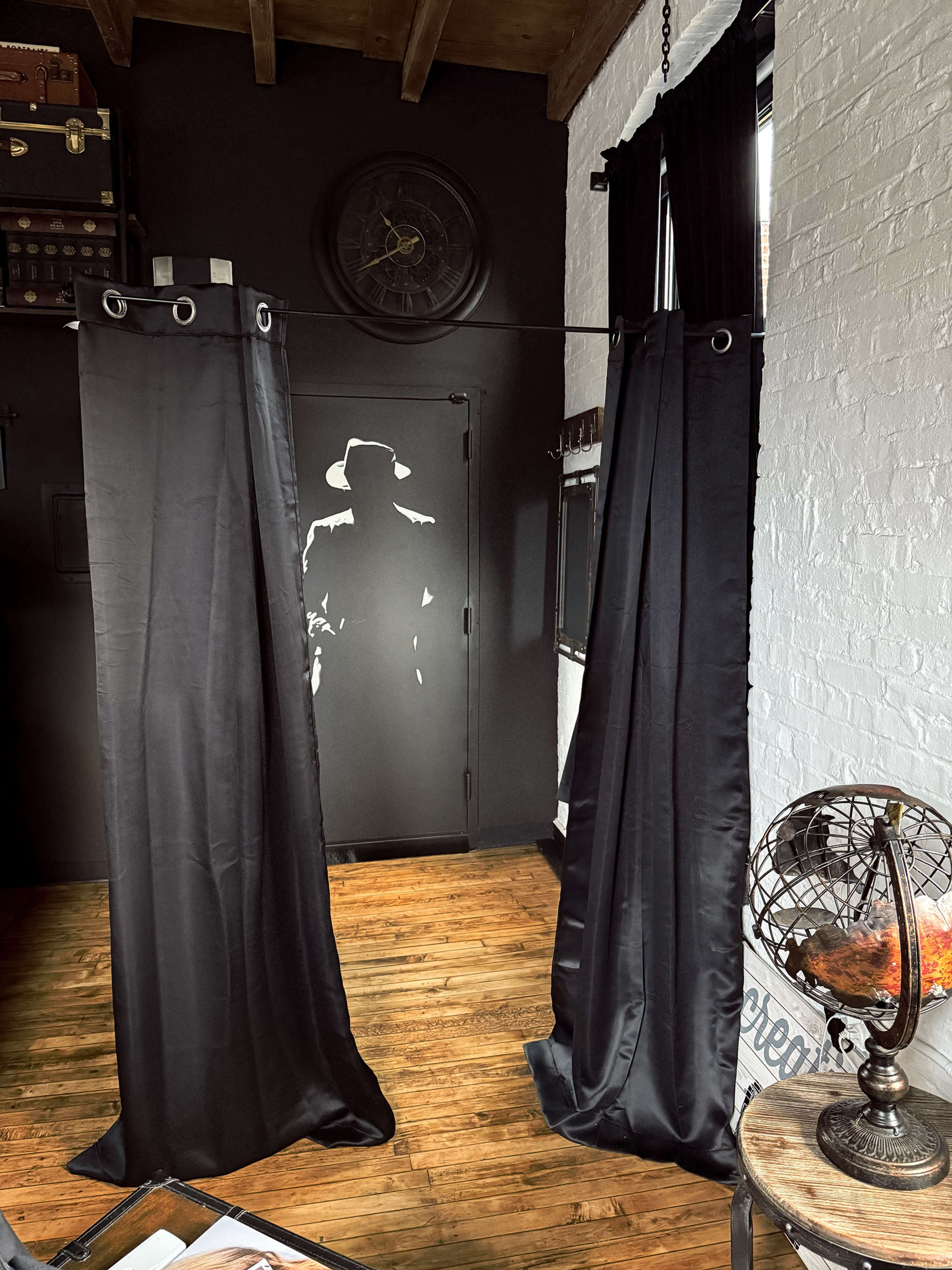 The image shows a room with wooden flooring, featuring two black fabric curtains hanging on either side, and a silhouette of a figure painted on the opposite wall.