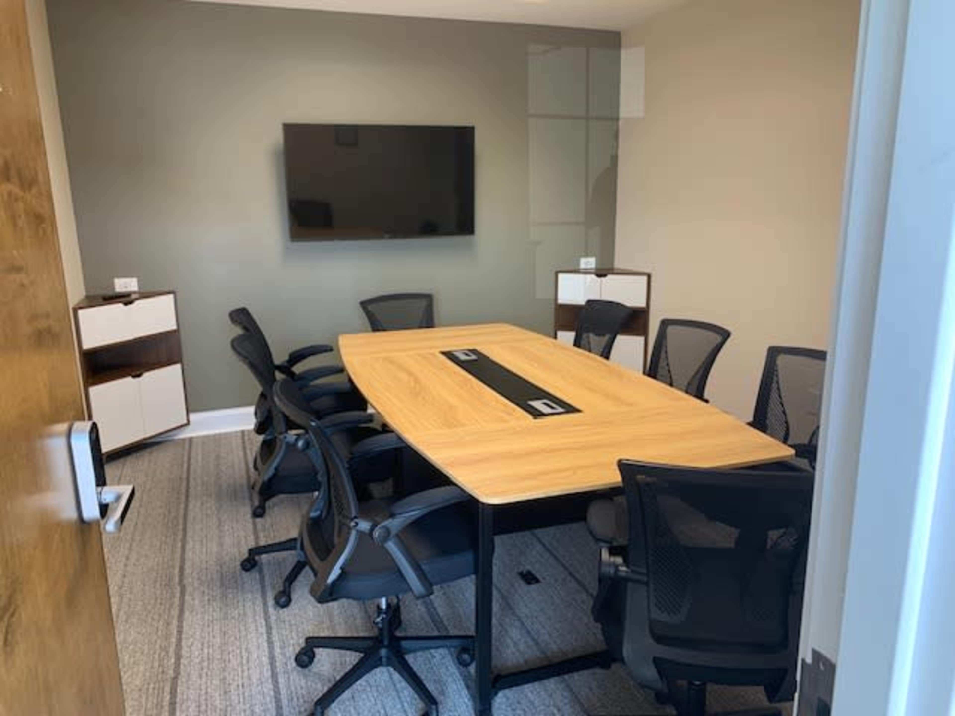 A modern conference room with a large wooden table surrounded by eight black chairs, a television mounted on the wall, and two cabinets in the corners.