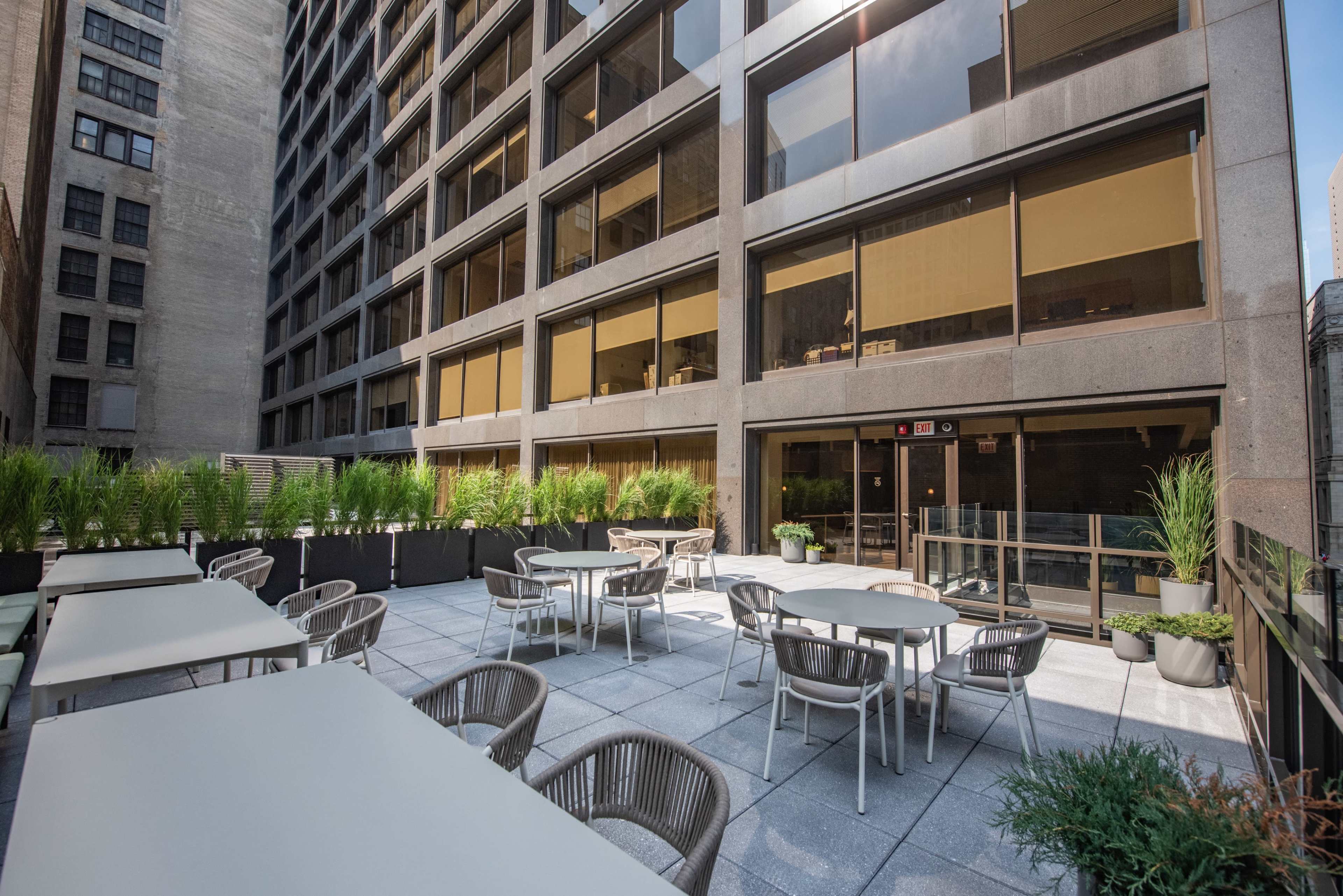 The image shows a rooftop patio with white tables and chairs surrounded by planters, adjacent to a modern building with large windows.
