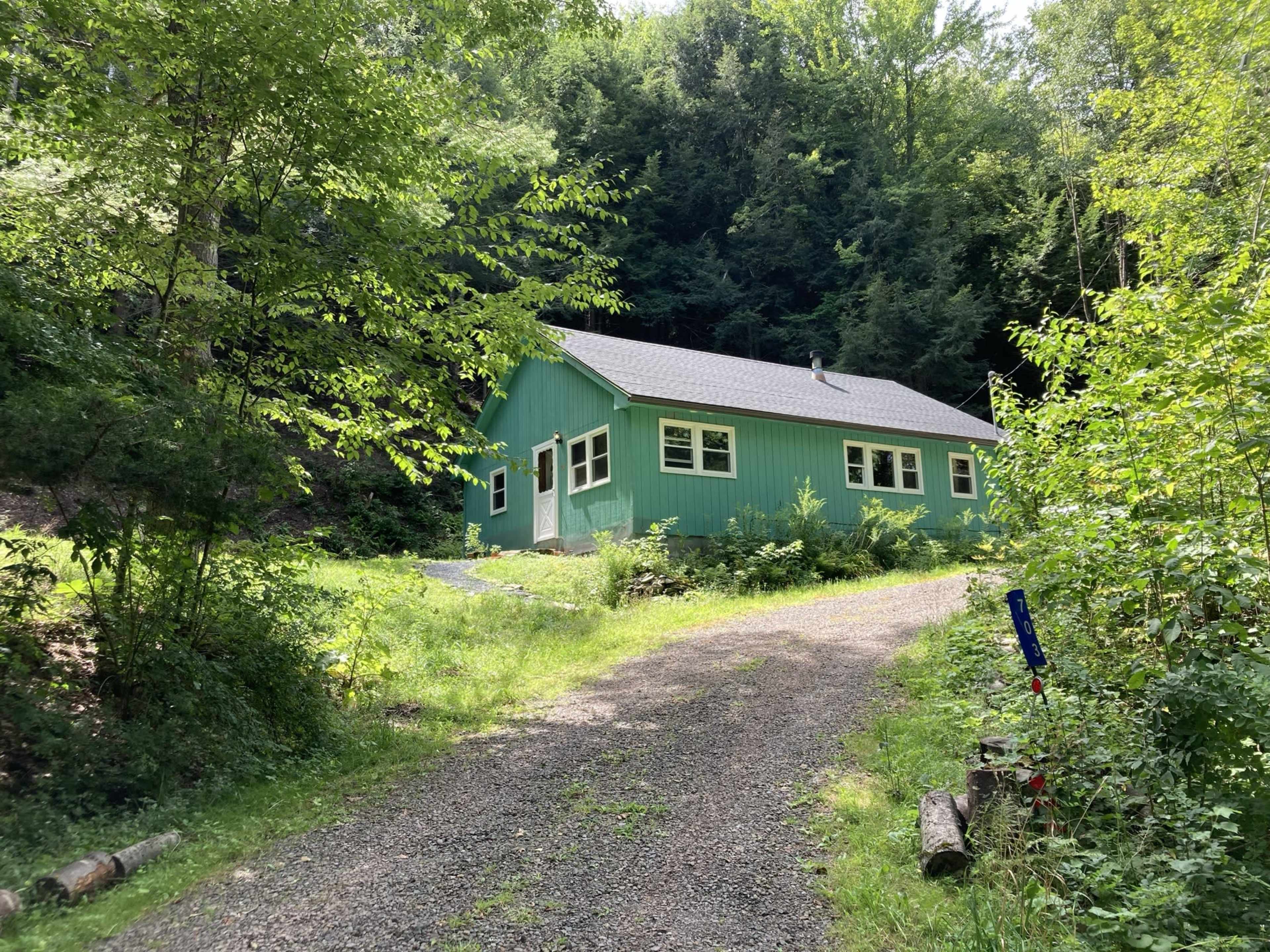 A green house is situated at the end of a gravel driveway, surrounded by lush greenery and trees.