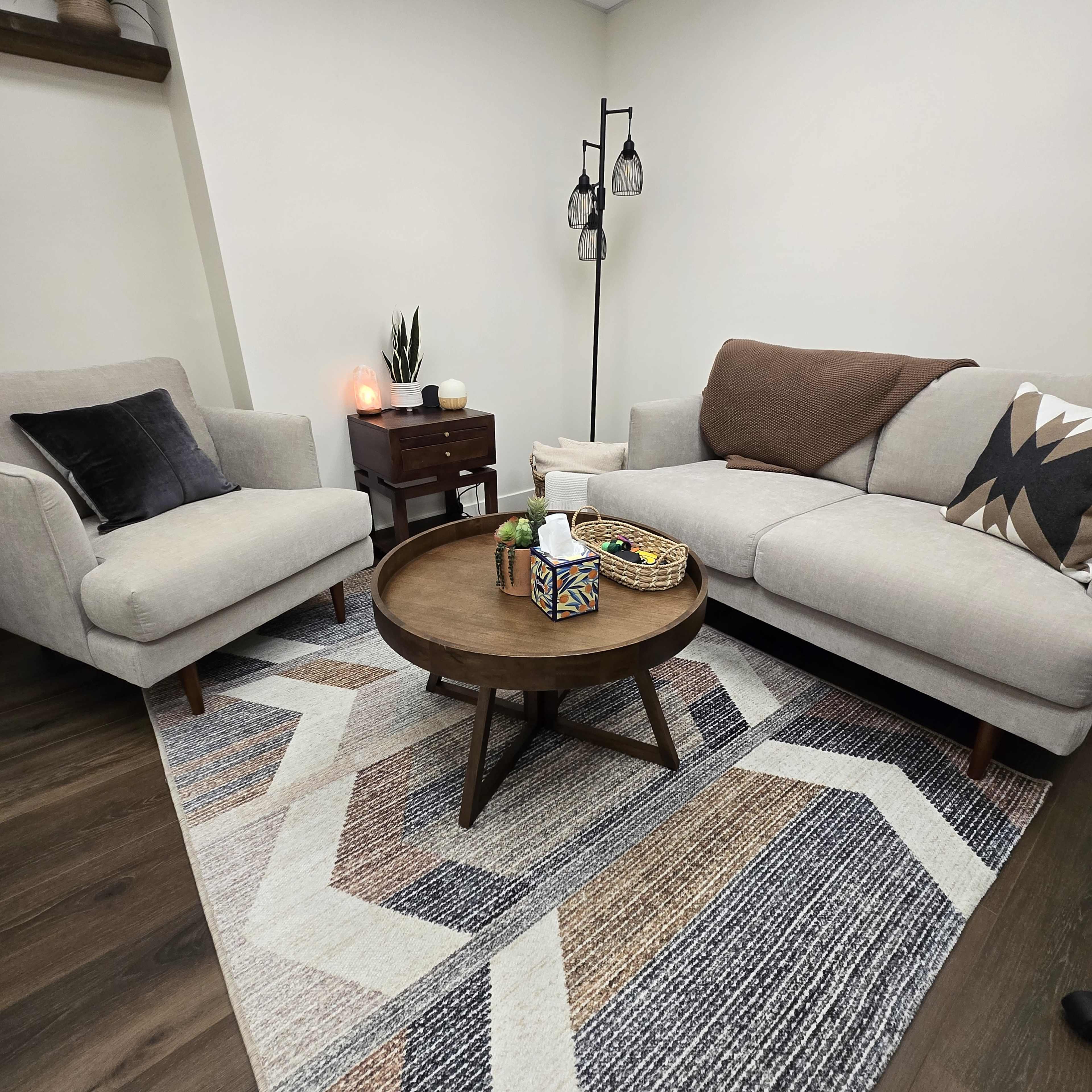 The image shows a cozy living room setup featuring a gray sofa, a light gray armchair, a round wooden coffee table, and a patterned area rug on a dark wooden floor.