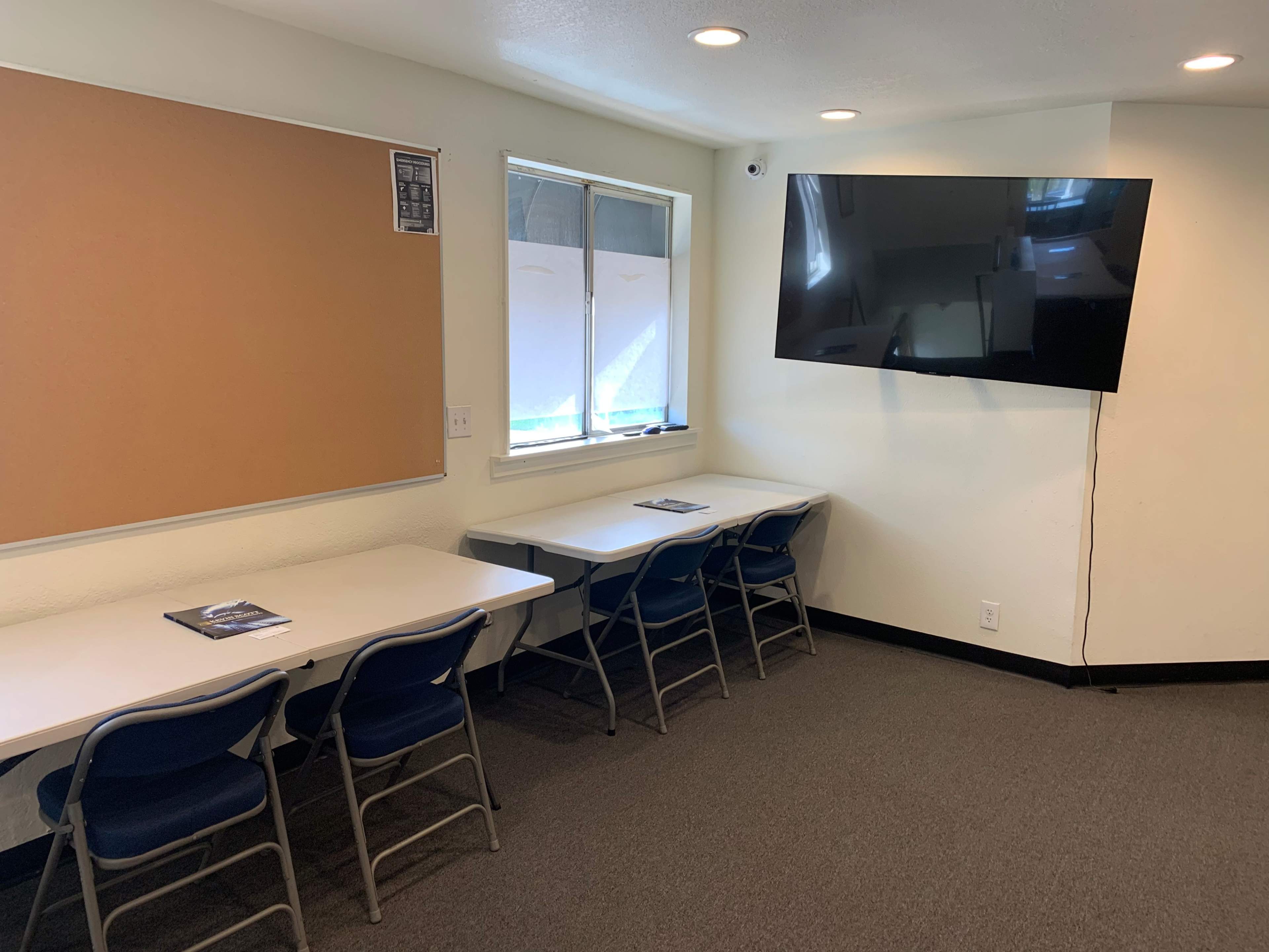 The room contains several white tables and blue chairs, a bulletin board, and a wall-mounted television.