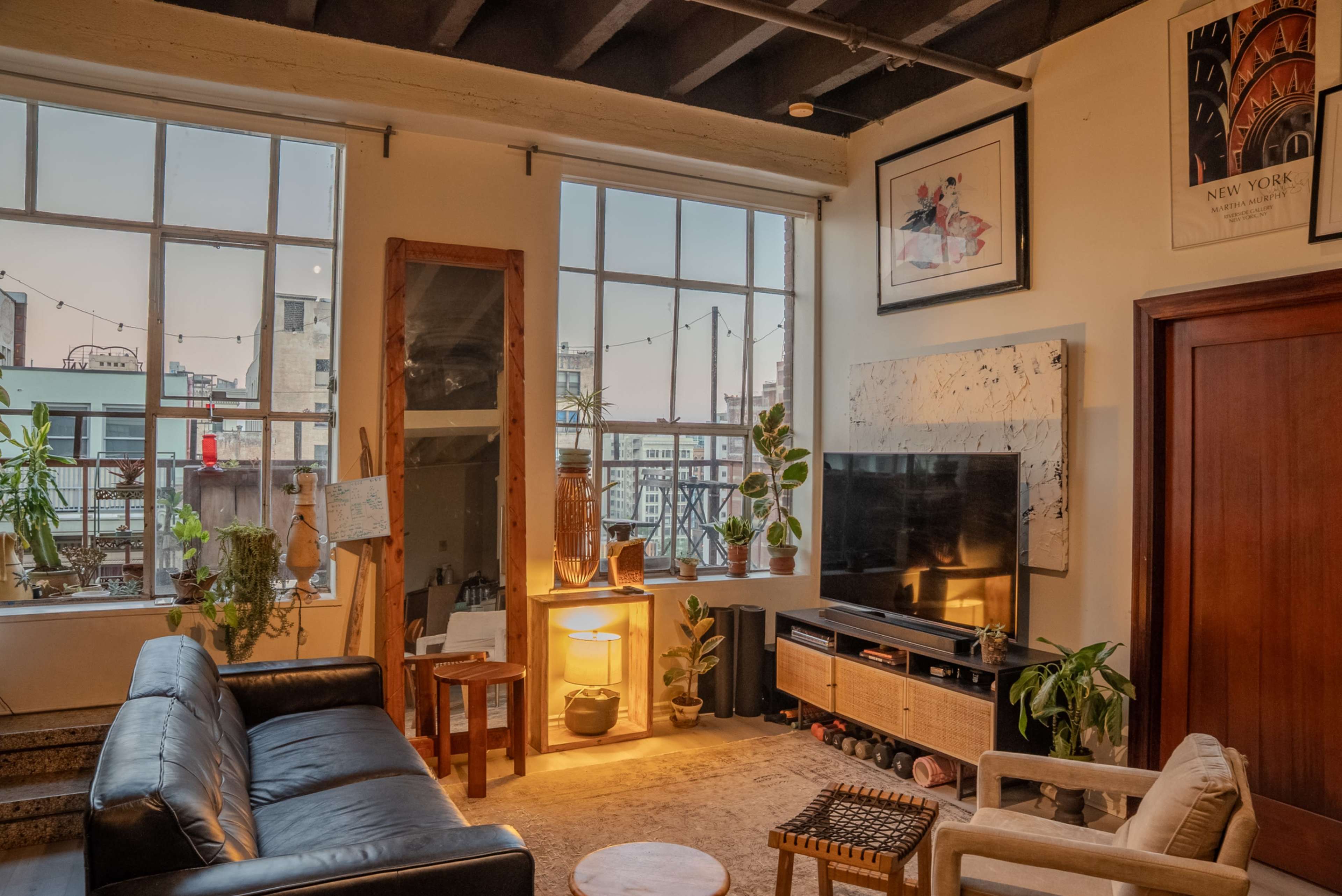 The living room features large windows, a black leather sofa, a wooden coffee table, and houseplants beside a television on a media console.