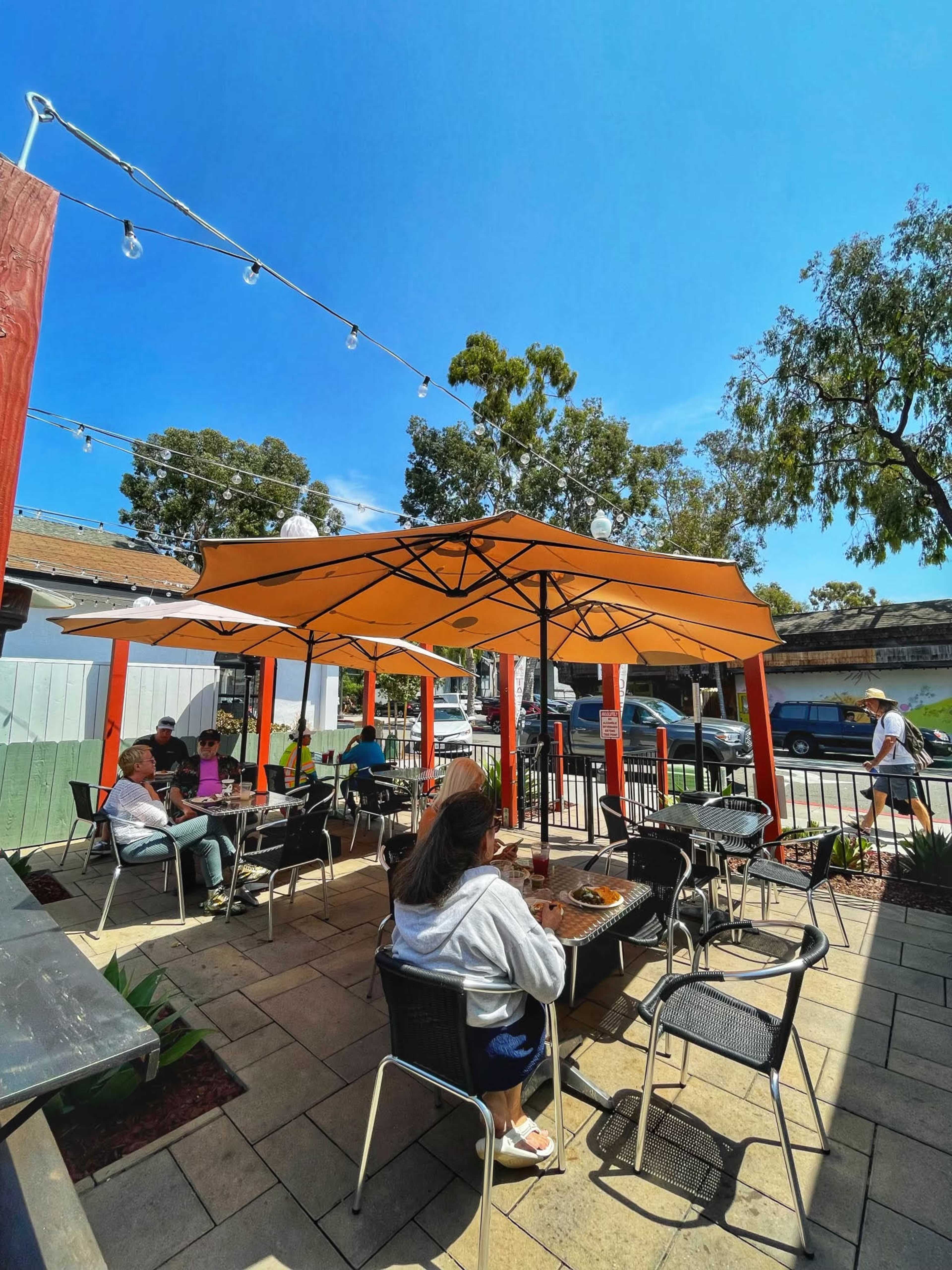 A patio with orange canopies features several people seated at tables, enjoying a sunny day.