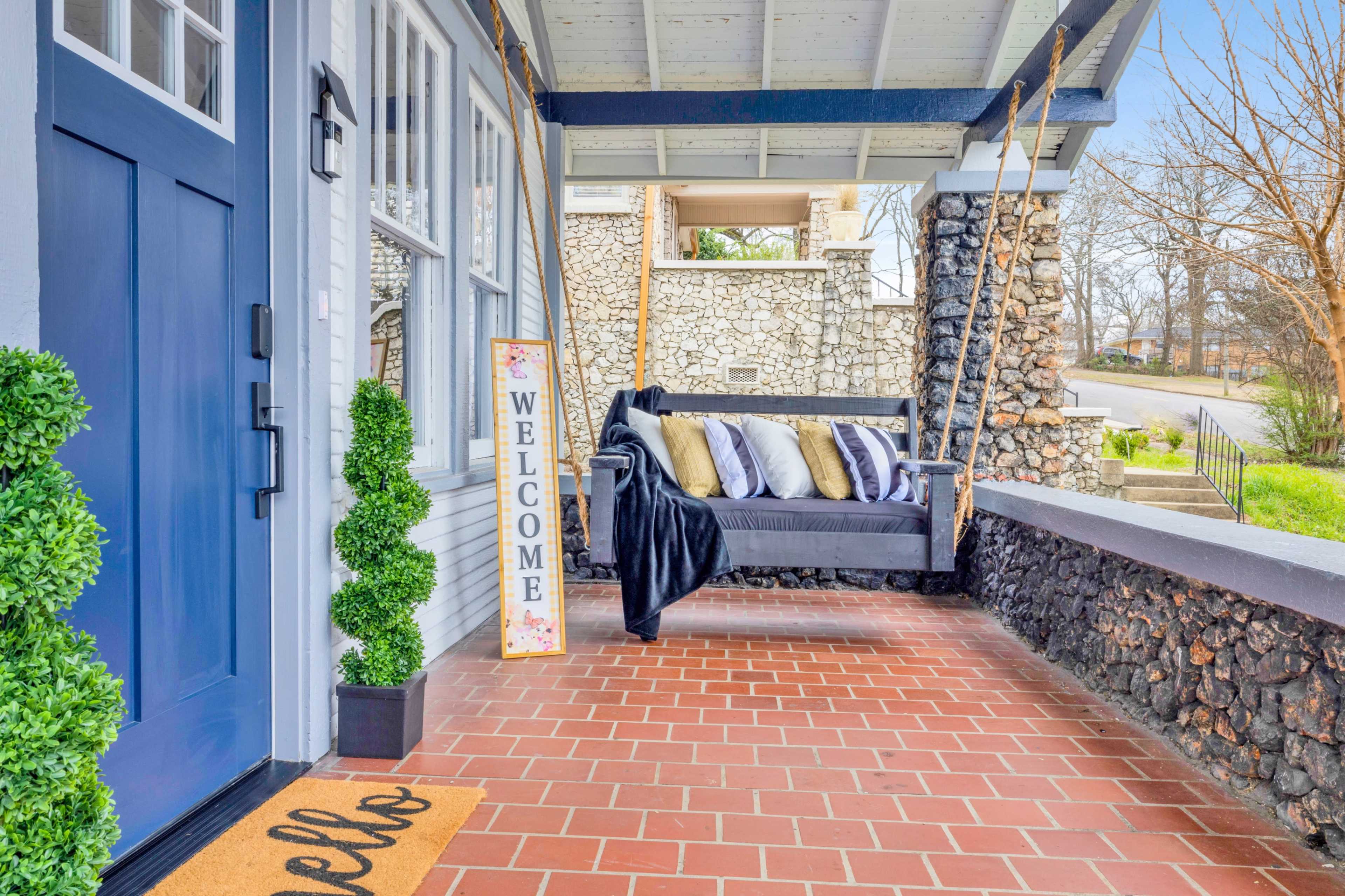 A porch with a blue front door, a welcome sign, a sofa adorned with pillows, a throw blanket, and potted plants.