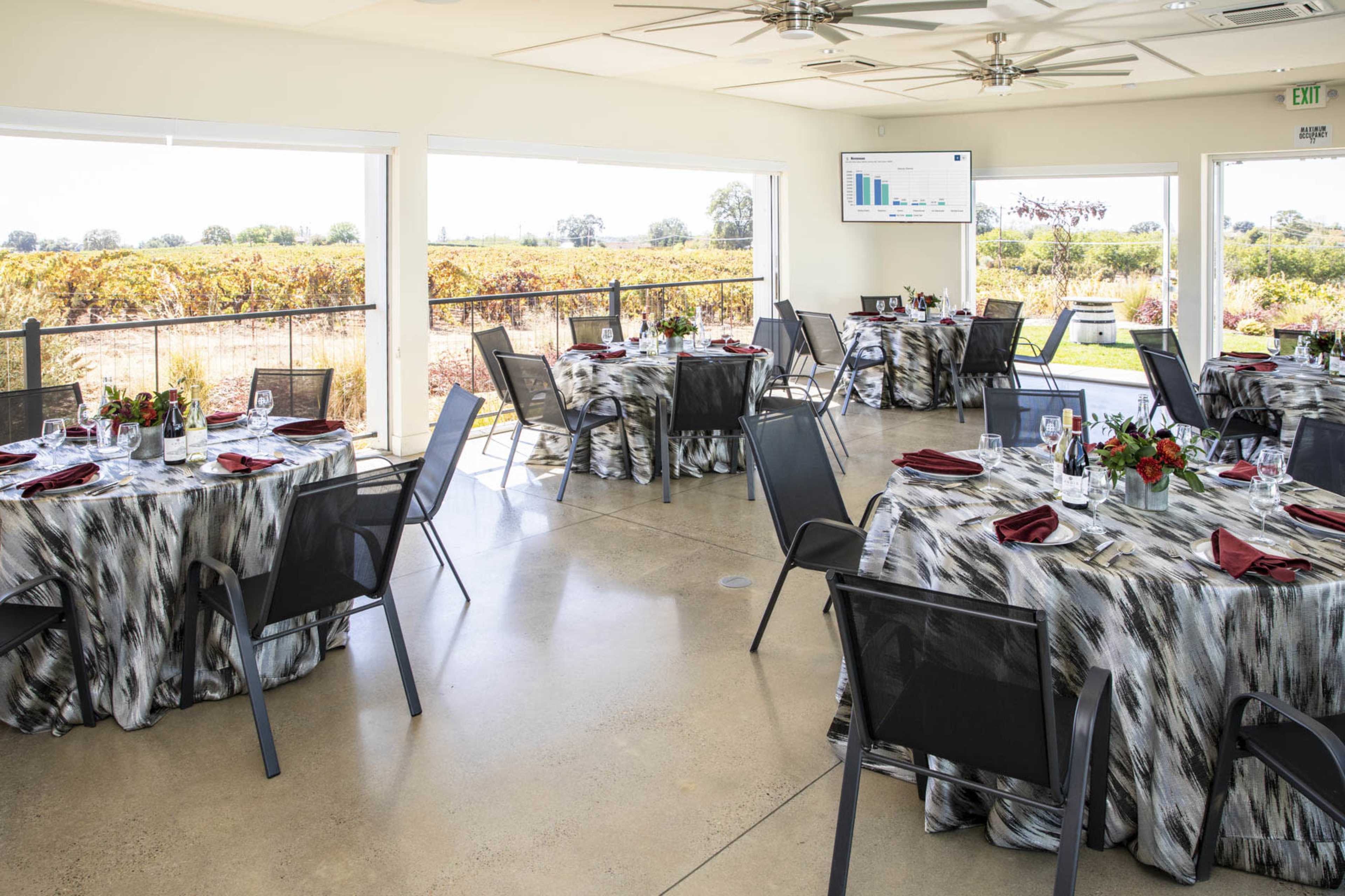 A dining area with several tables set for a meal, featuring tablecloths and chairs, surrounded by large windows overlooking a vineyard.