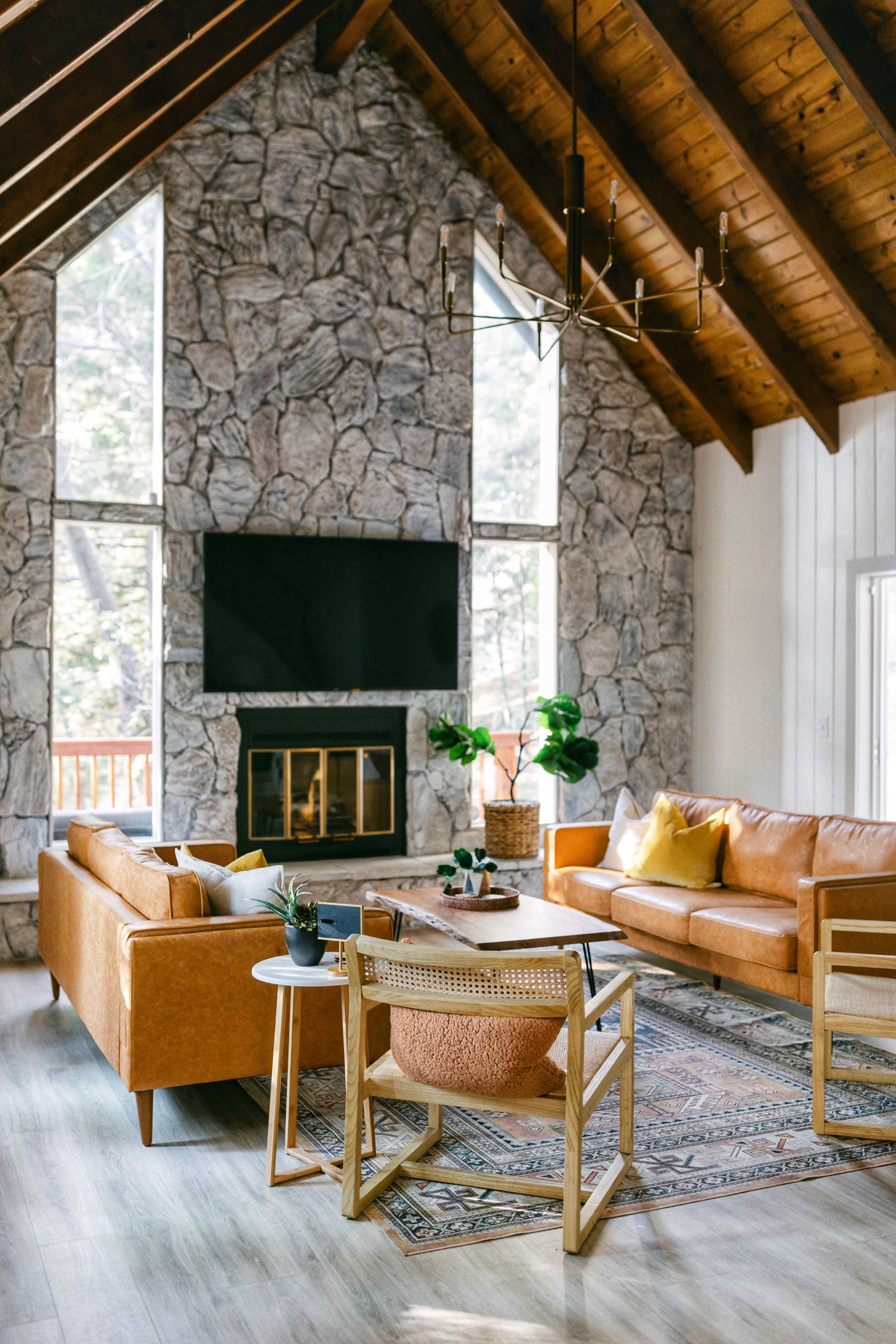 The image depicts a cozy living room with a high stone wall, large windows, a flat-screen TV above a fireplace, and leather sofas arranged around a decorative rug.