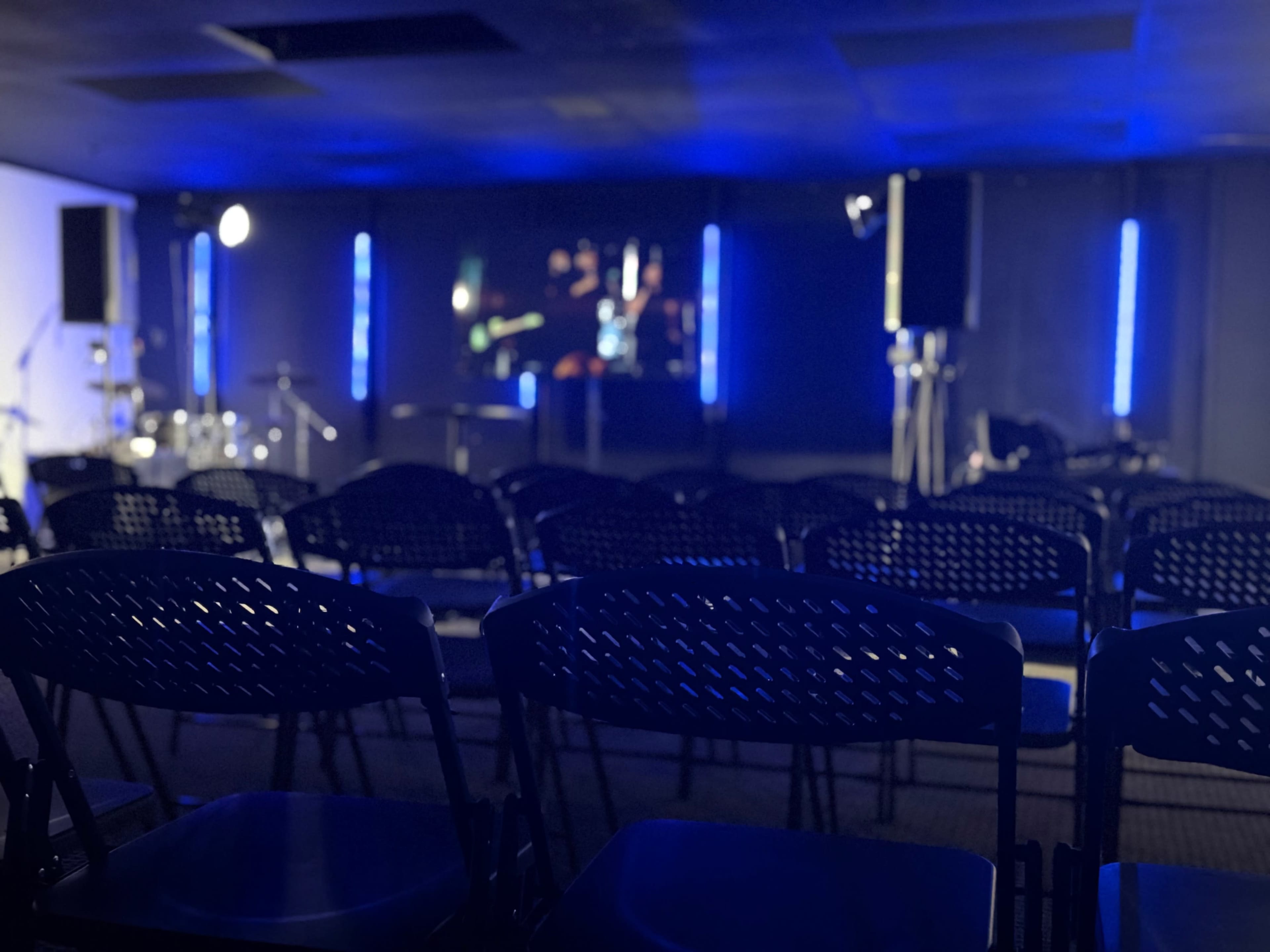 The image shows a row of black chairs facing a stage with blue lighting and a screen displaying visuals.