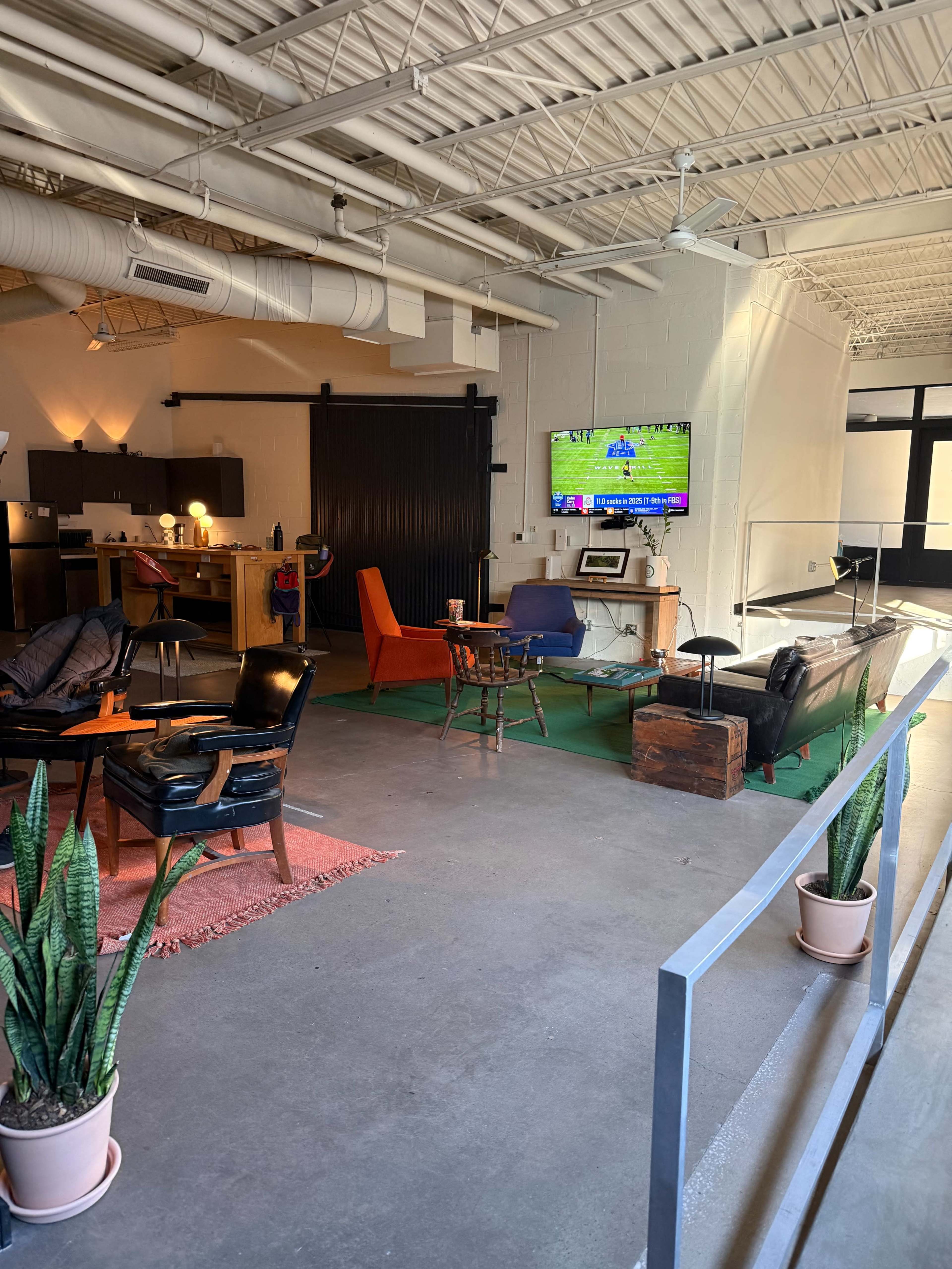 The image shows a spacious, modern lounge area with various seating options, a television mounted on the wall, and potted plants.