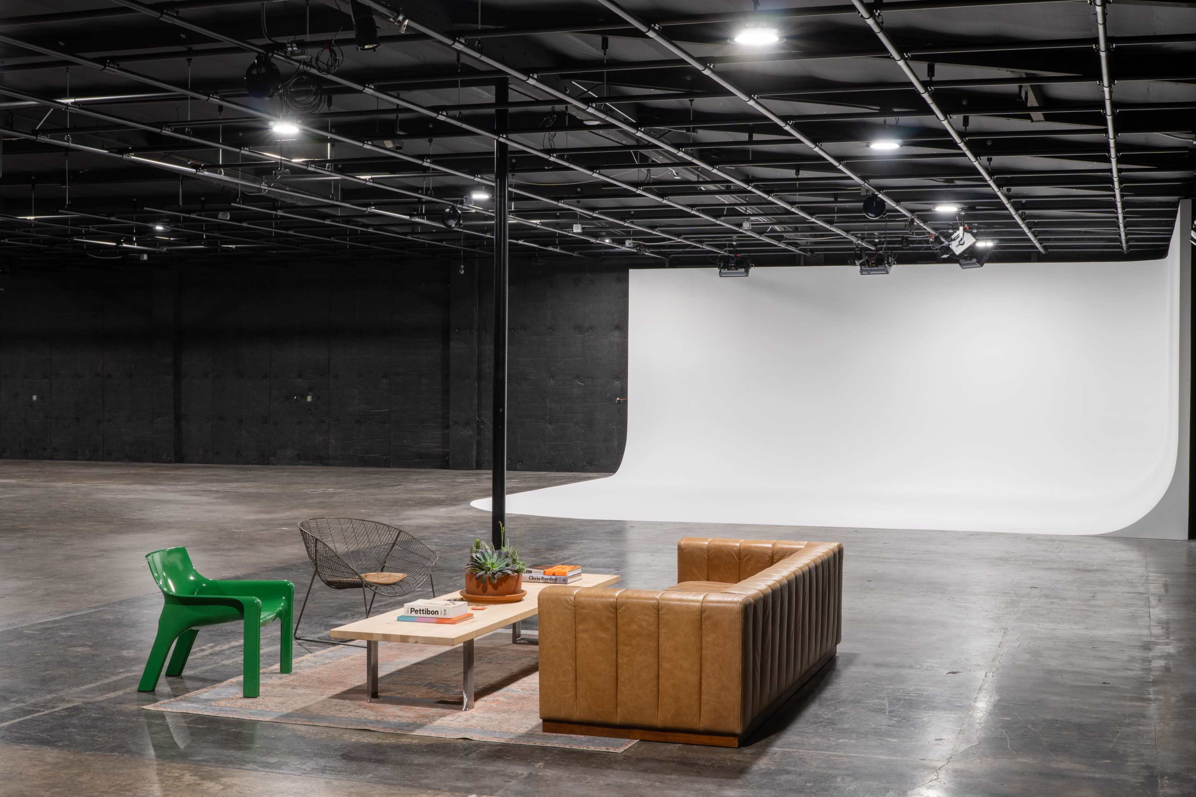 A spacious studio with a minimalist wooden couch, a coffee table, a green chair, and a white backdrop for photography.