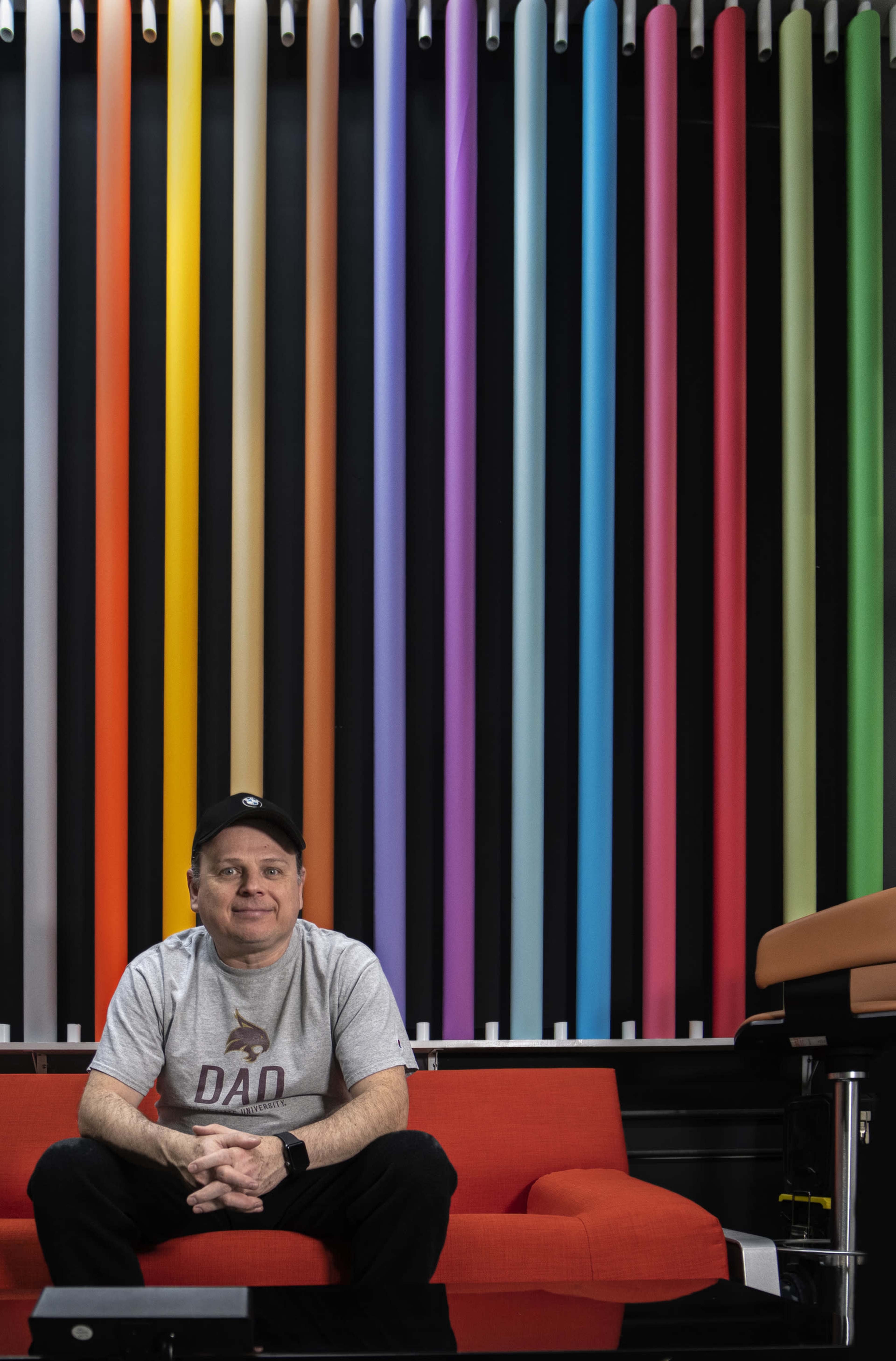 A man sits on a red couch in front of a wall adorned with colorful vertical tubes.