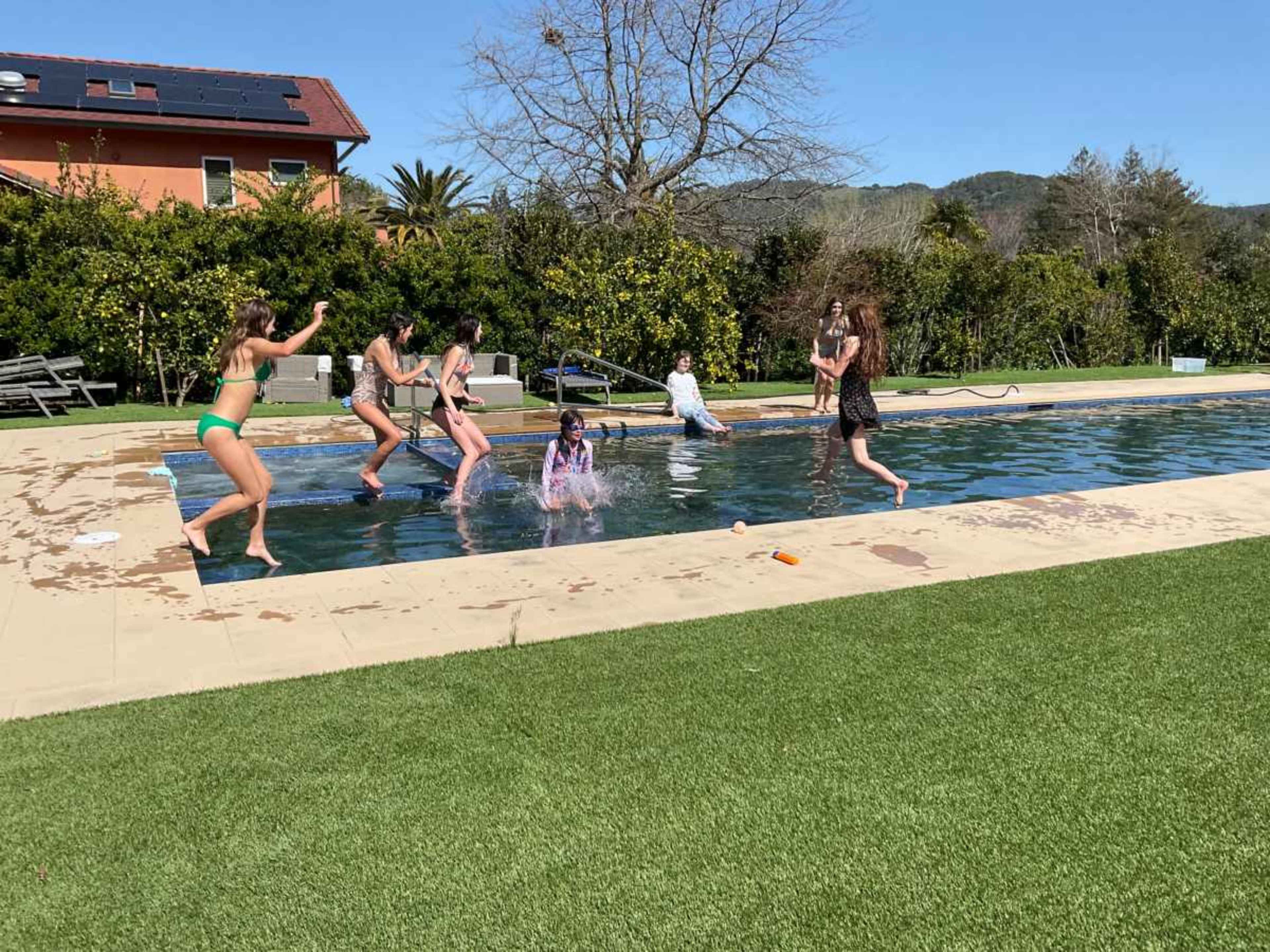 A group of six people is playing and splashing in a swimming pool surrounded by greenery on a sunny day.