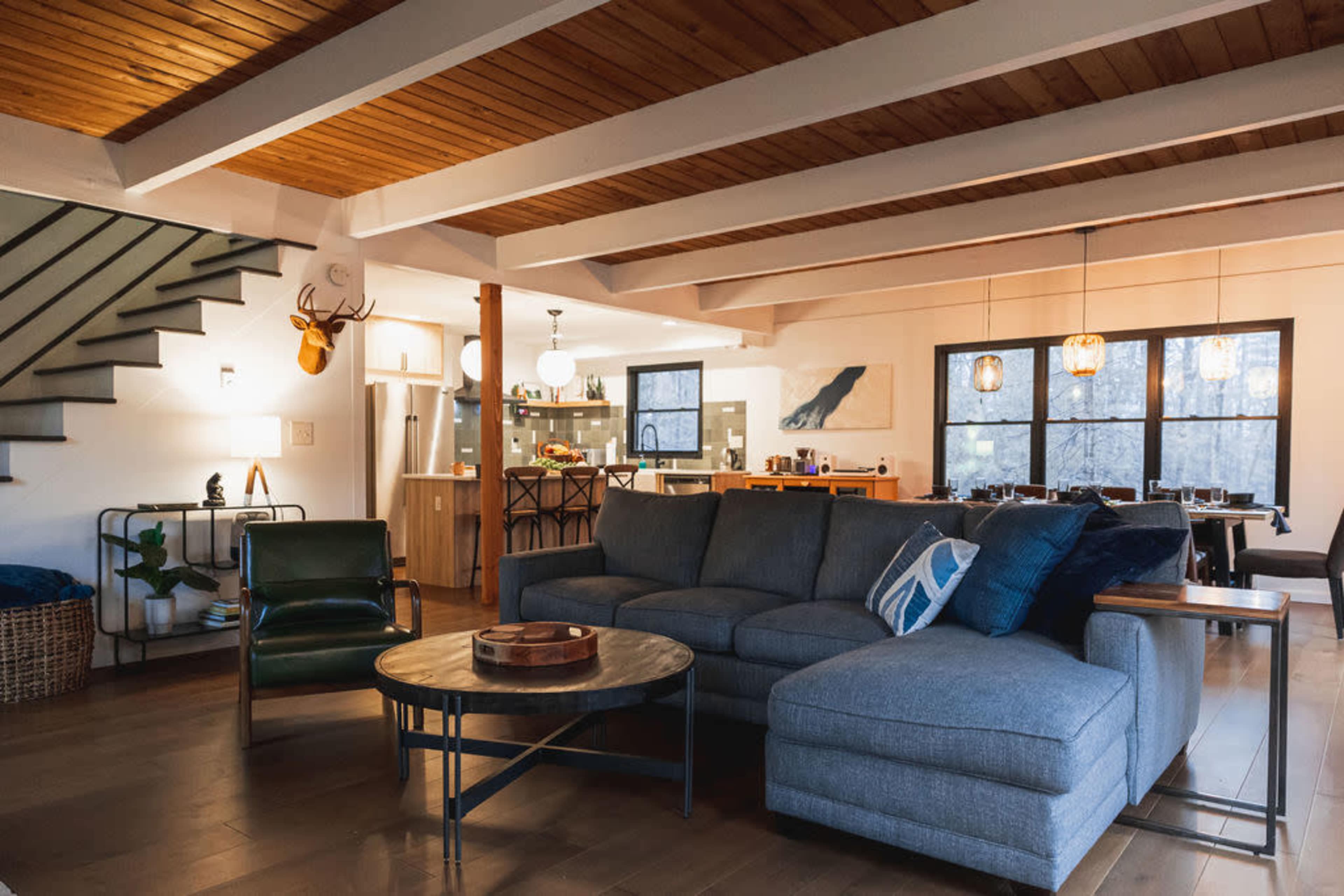 The image shows a modern living room with a large sectional sofa, a coffee table, and an open layout connecting to a kitchen area.