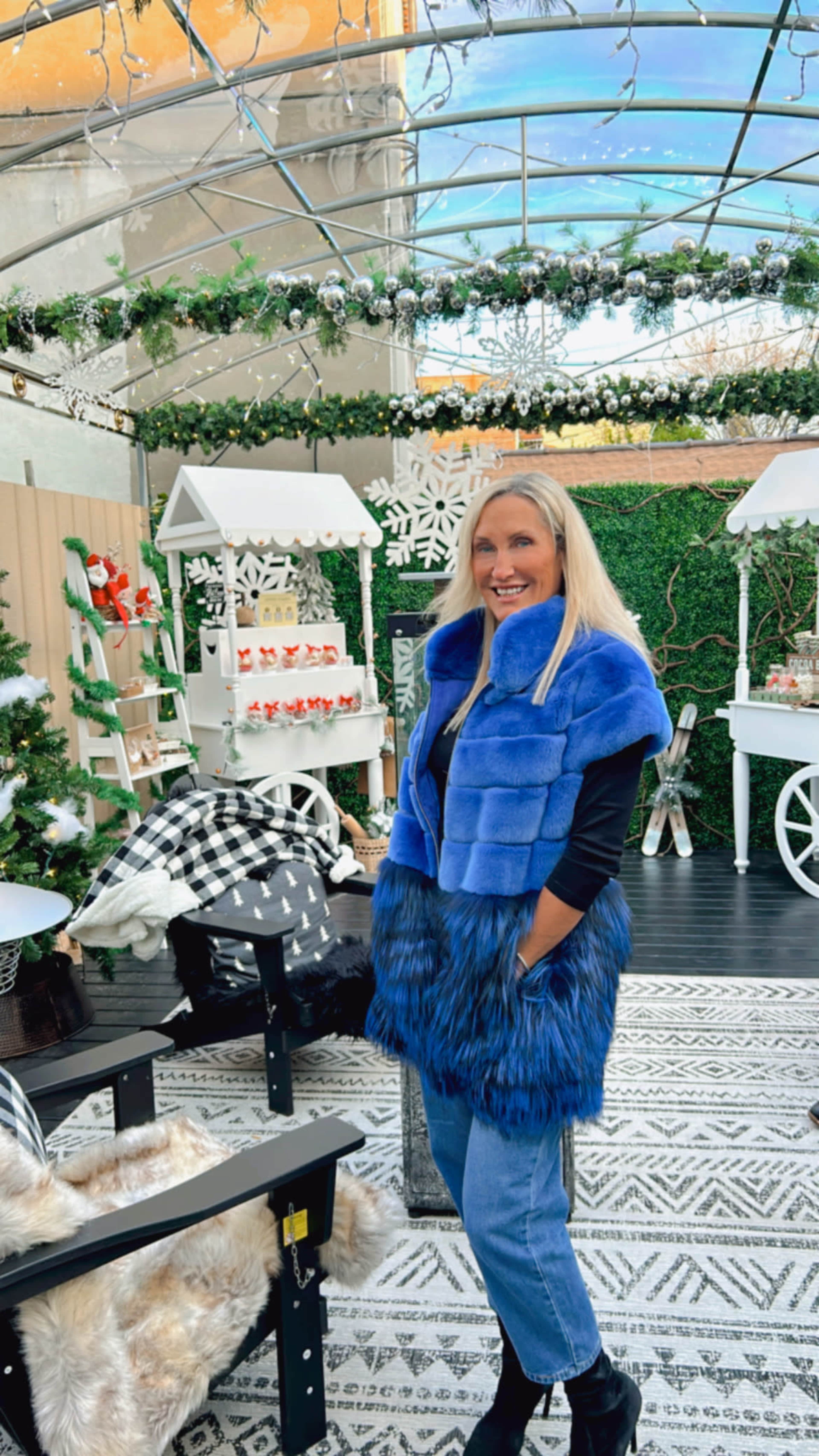 A woman in a blue fur vest stands in a decorated indoor space with holiday-themed displays and greenery.