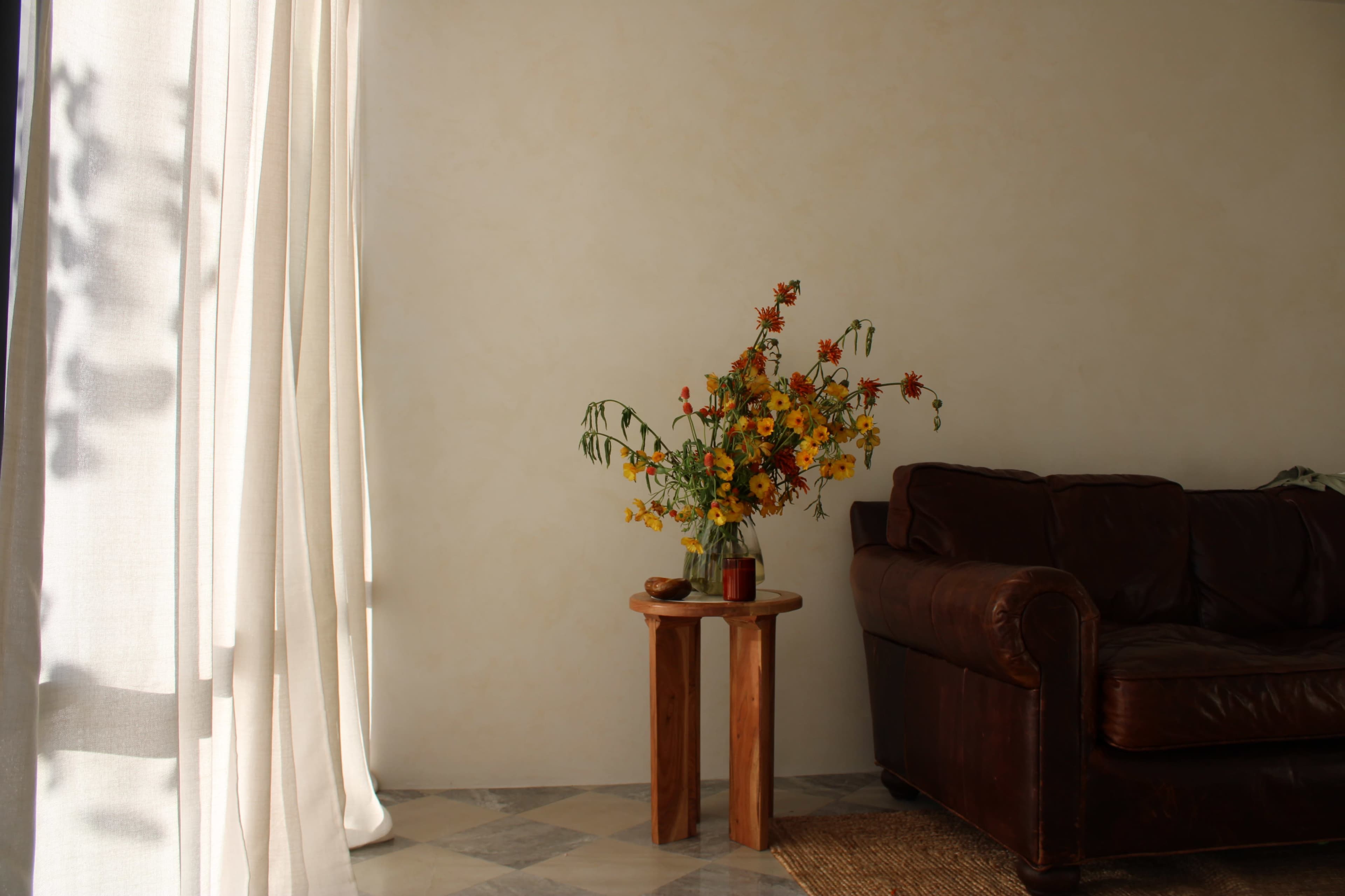 A vase of flowers sits on a wooden table next to a brown leather couch in a room with natural light coming through sheer curtains.