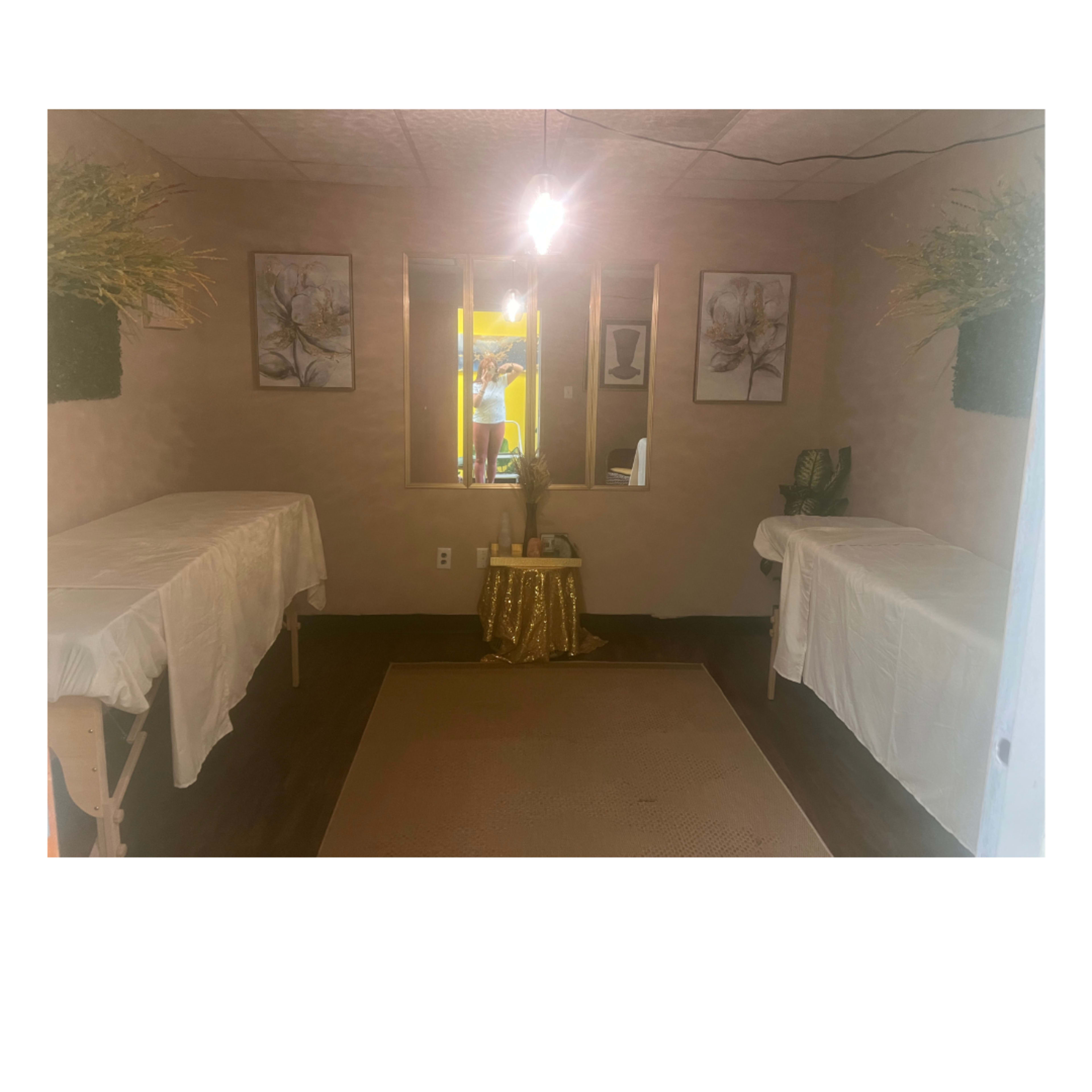 The image shows a small therapy room with two massage tables, a decorated table in the center, and framed artwork on the walls.
