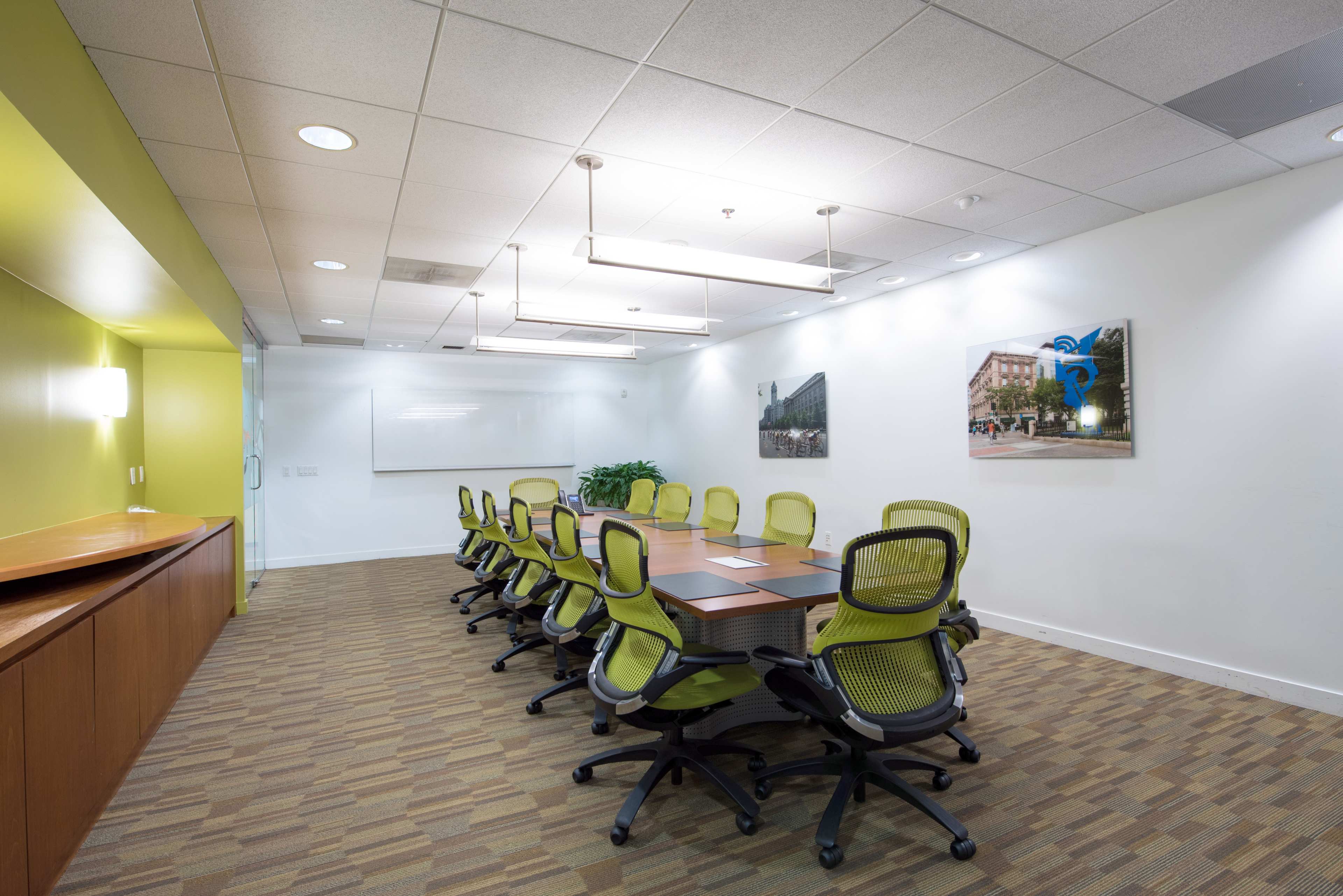A conference room is set up with a long wooden table surrounded by ergonomic chairs, featuring large windows and artwork on the walls.