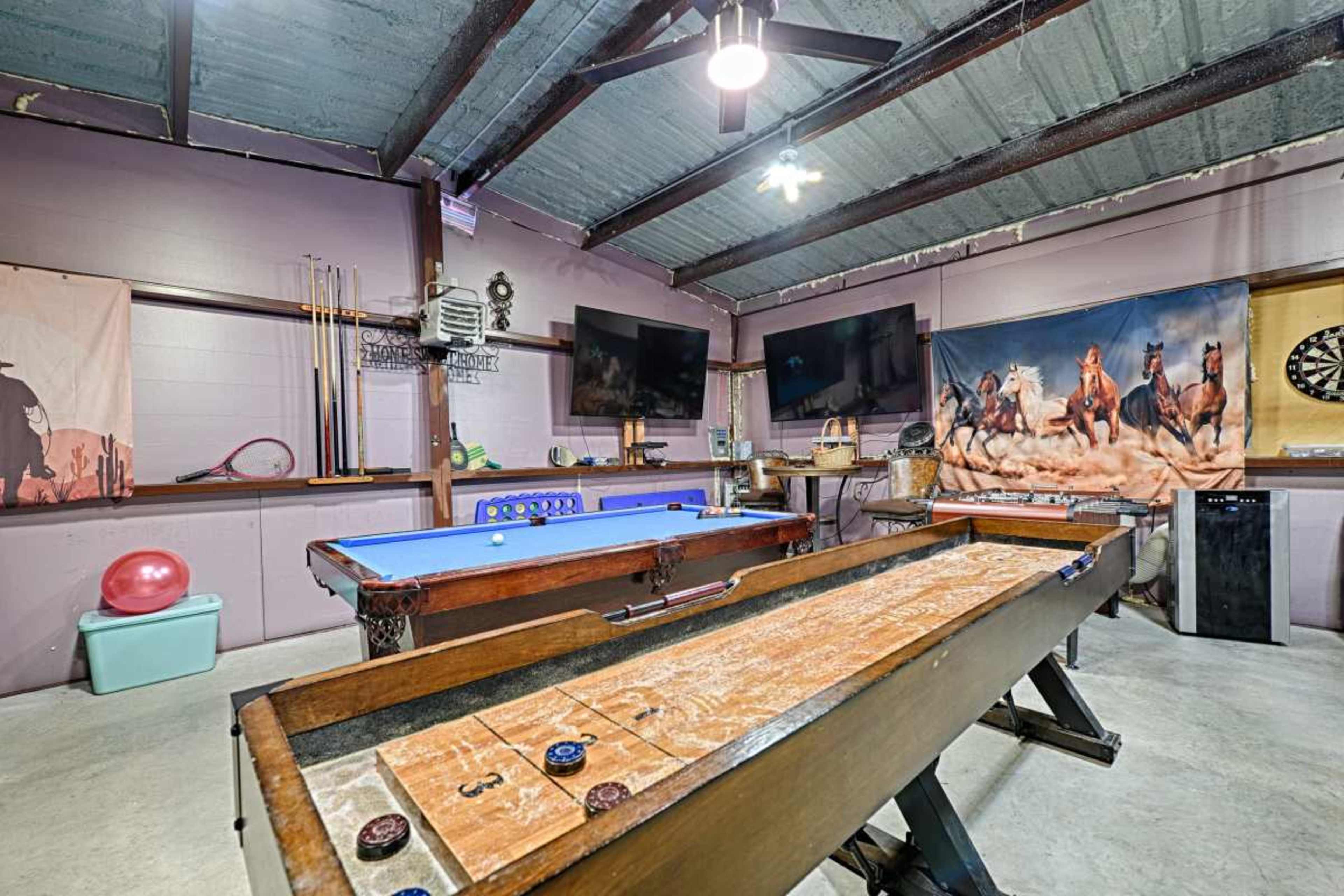 The image shows a recreation room with a blue pool table, a shuffleboard table, two mounted televisions, and various sporting equipment on display.