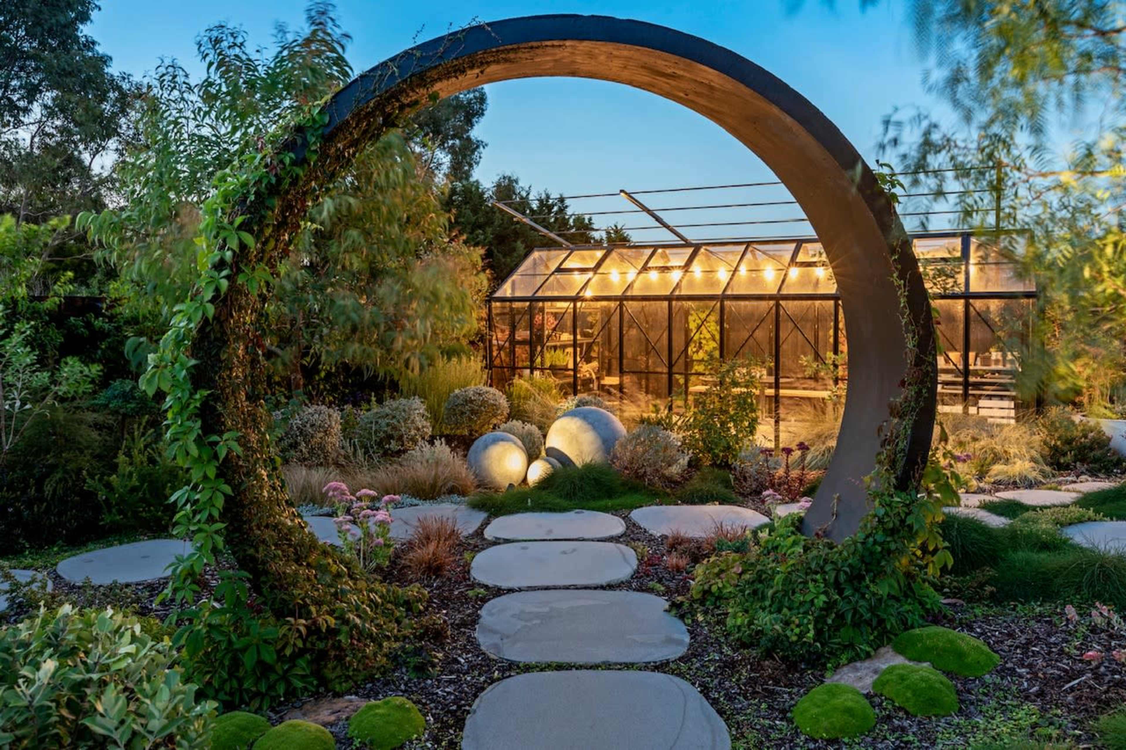 A circular stone archway frames a garden path leading to a greenhouse illuminated by nighttime lights.