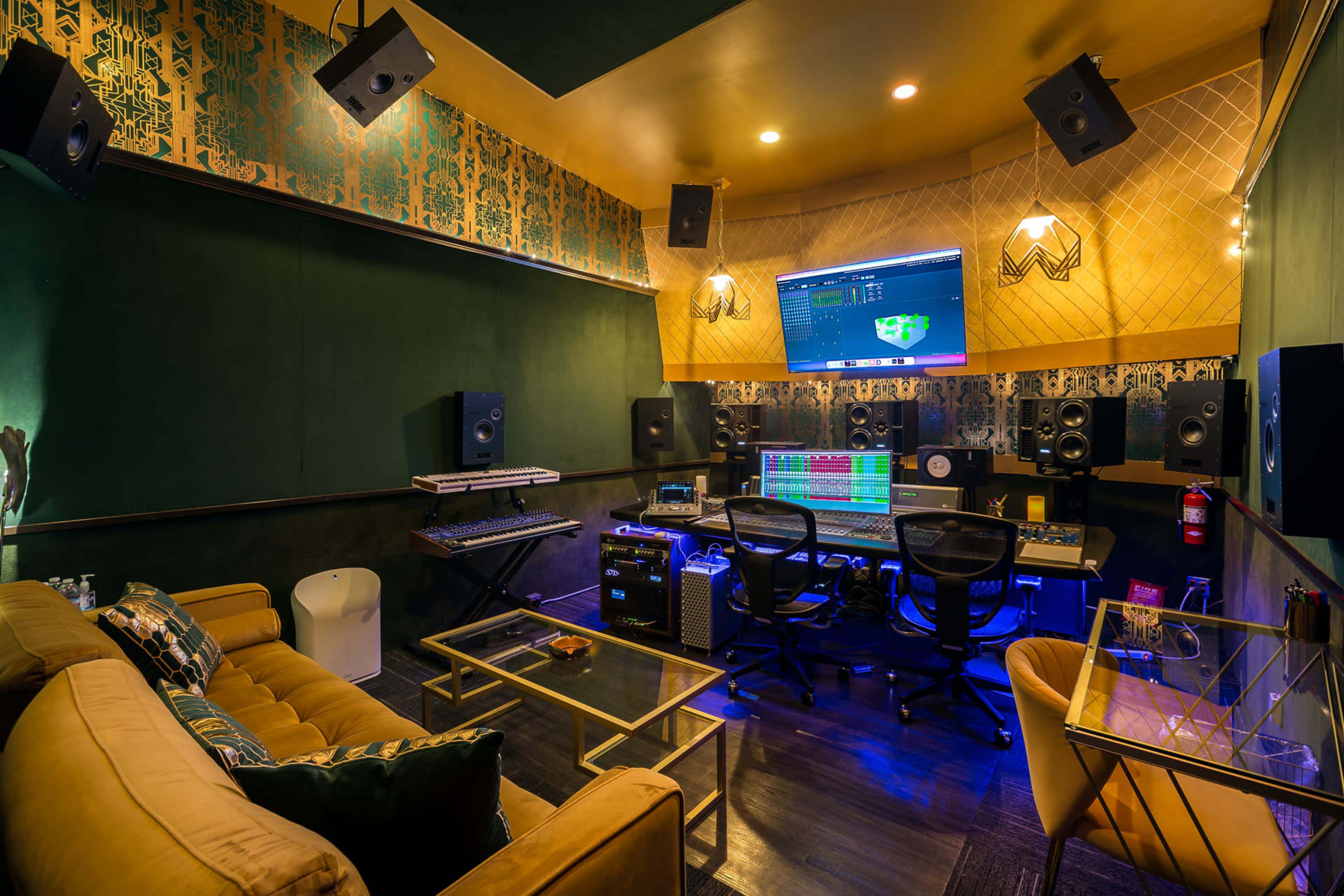 A sound recording studio features a large mixing console, multiple speakers, and a computer display, surrounded by green wallpaper and comfortable seating.