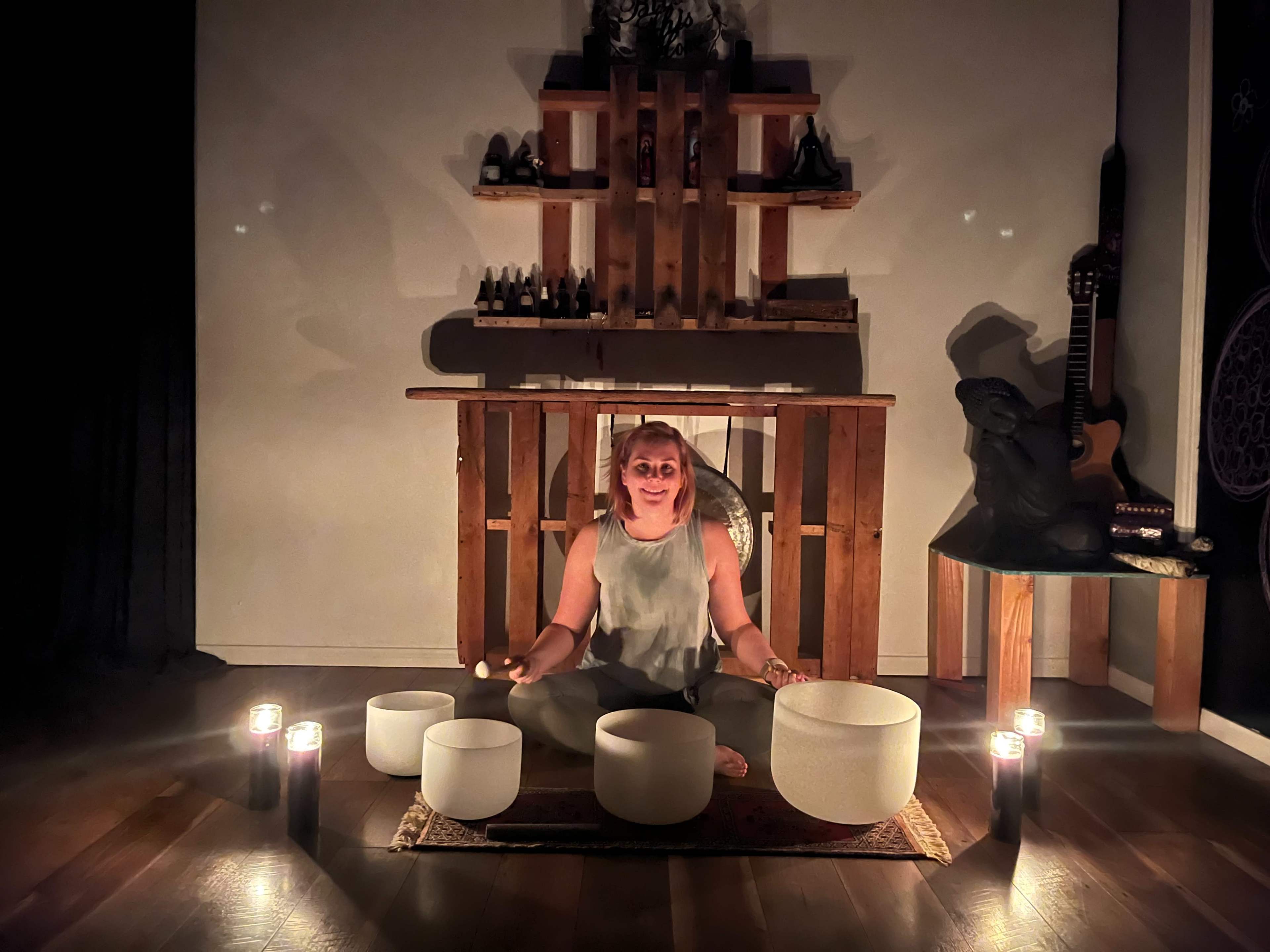 A person sits cross-legged on a rug surrounded by several crystal singing bowls and lit candles in a dimly lit room.